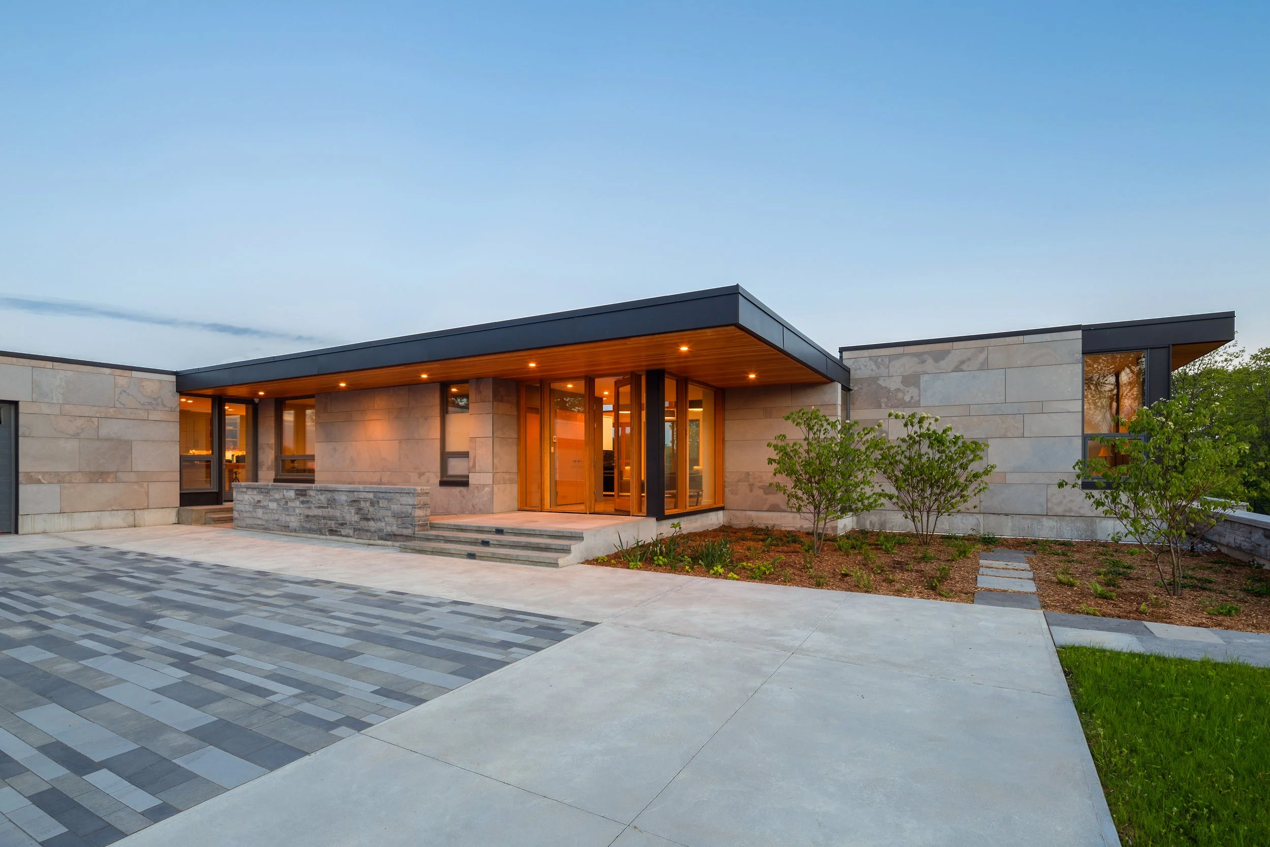 Modern house with stone and glass exterior, driveway, and landscaped garden with trees, under a clear evening sky.