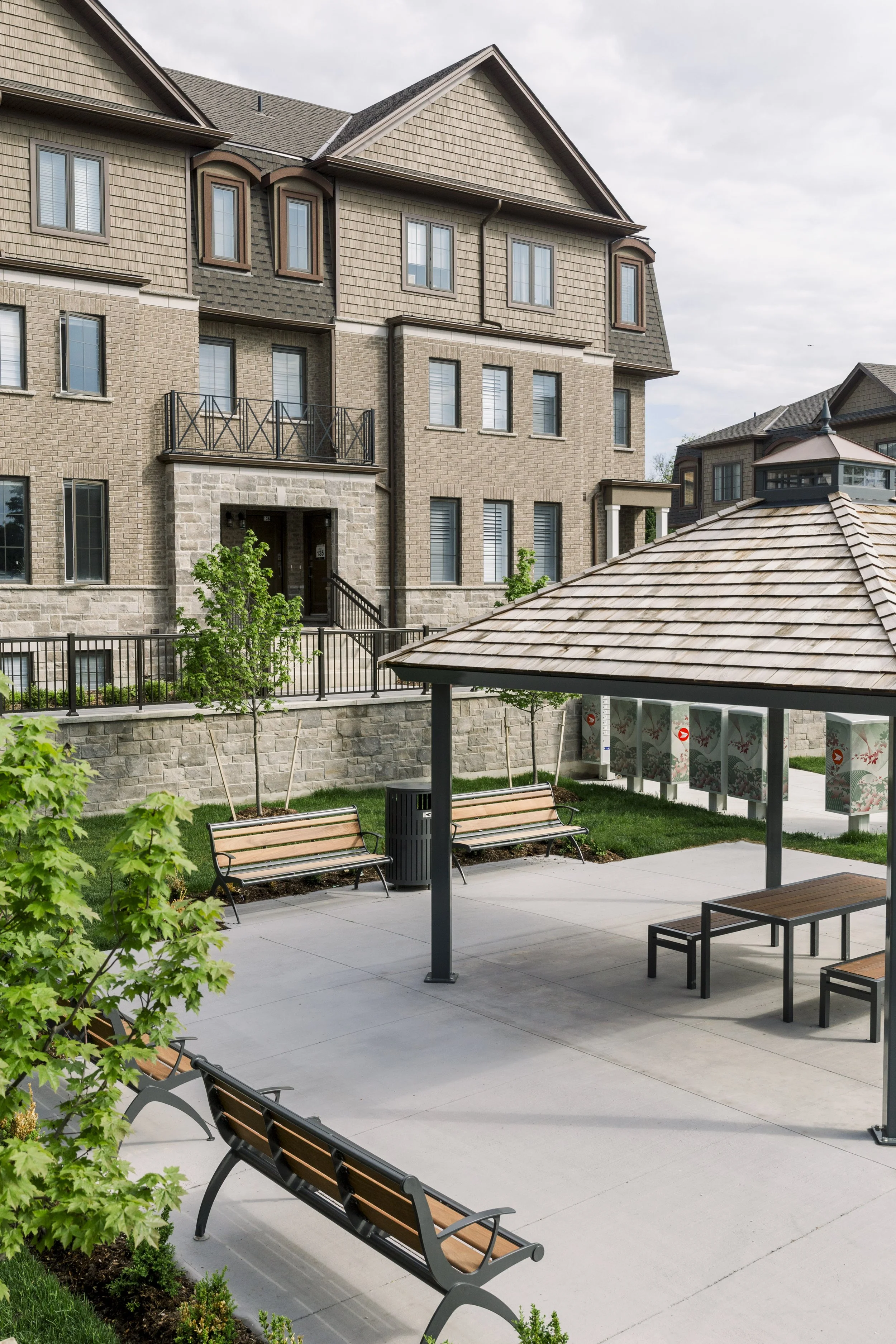 A small park area in front of a multi-story apartment building, with wooden benches, a trash can, a small pavilion with a shingled roof, and young trees along the sidewalk.