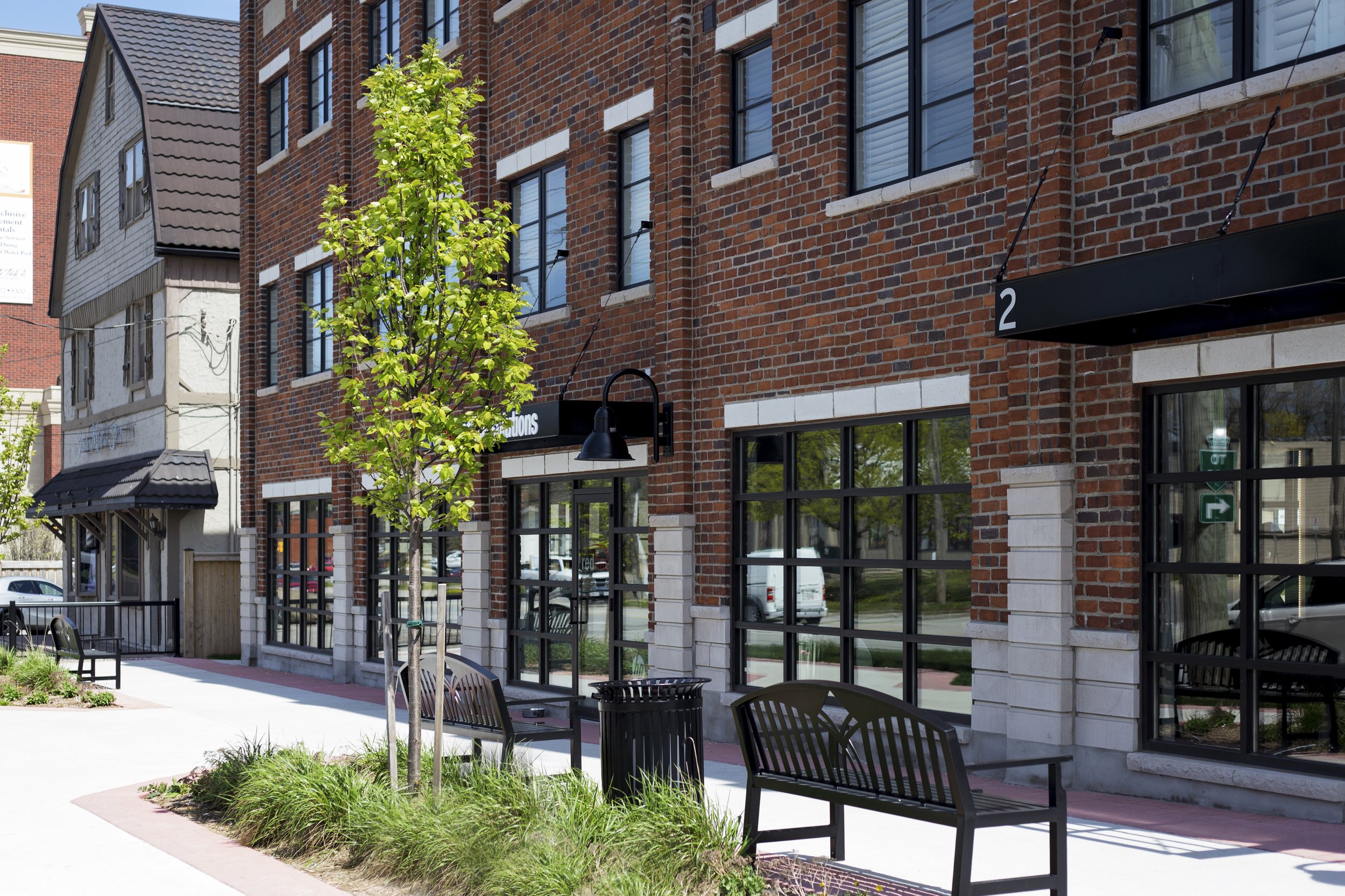 Street view of a brick building with large windows, a small tree, benches, and a trash can outside.