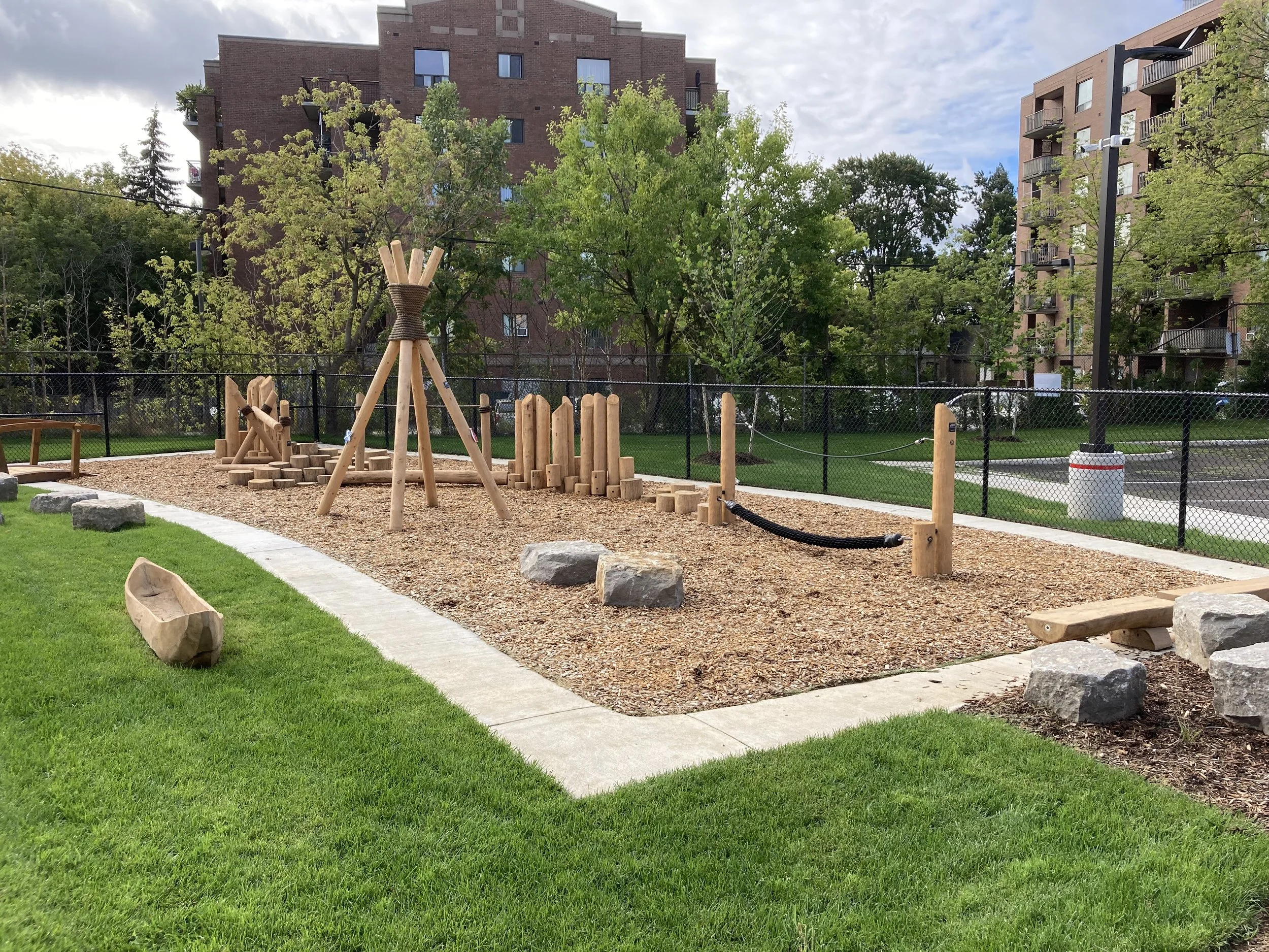 A playground with wooden climbing and balancing equipment, large rocks, and a green lawn, surrounded by a black metal fence and residential apartment buildings in the background.