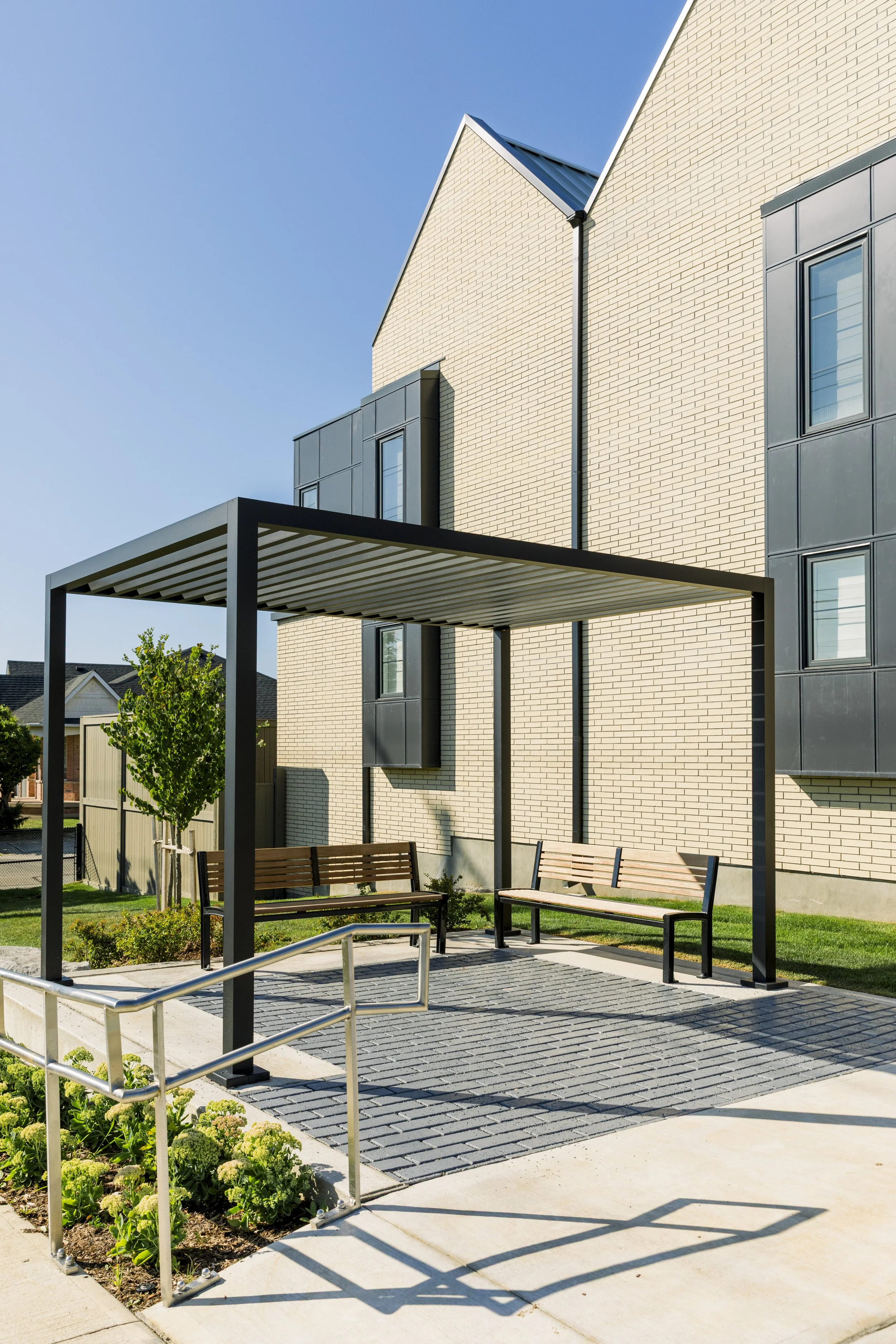 Modern outdoor seating area with two benches under a metal pergola, adjacent to a contemporary brick housing unit with black window frames, small garden with greenery, and a paved walkway.