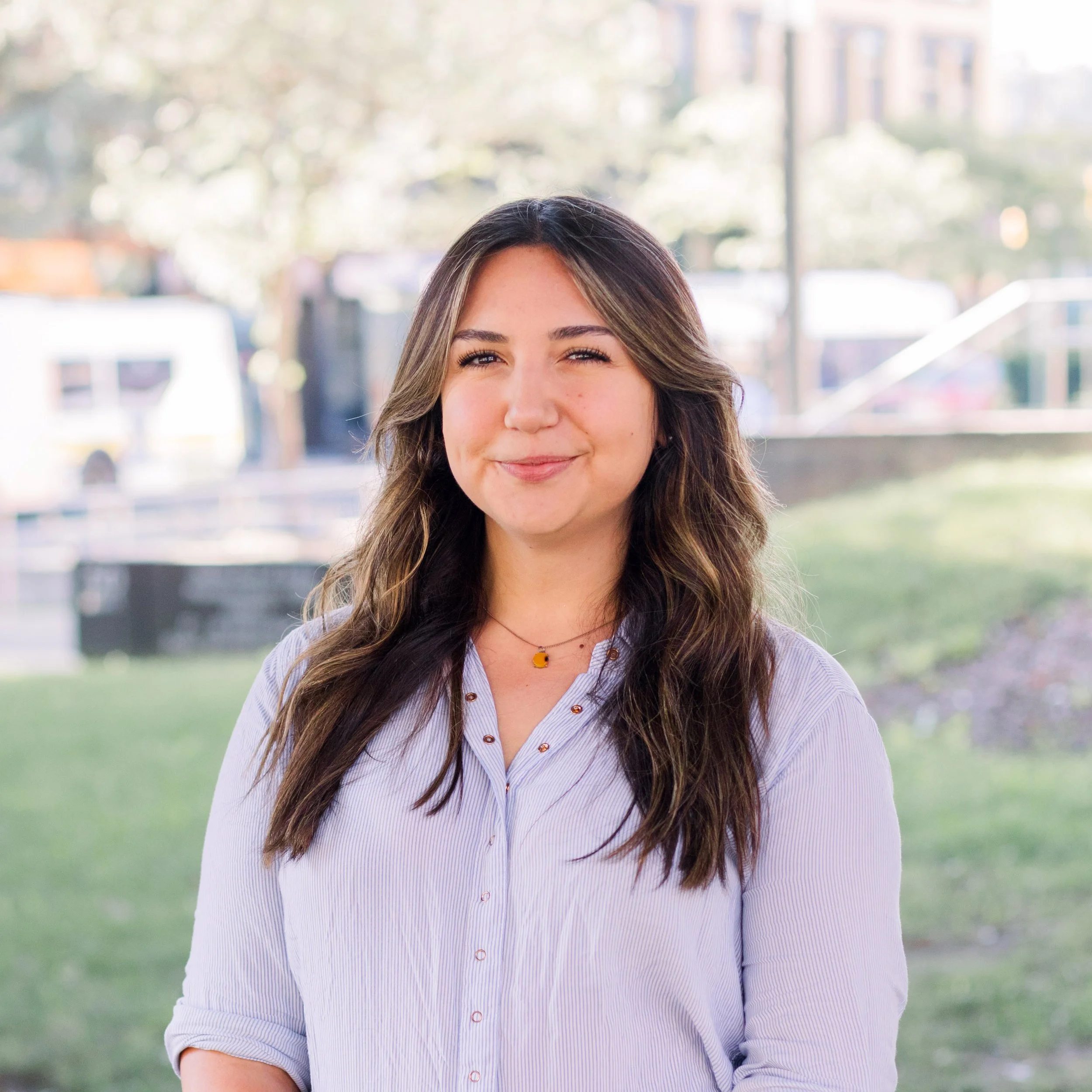 A young woman with long brown hair smiling outdoors, wearing a light purple button-up shirt and a small necklace, with a blurred background of greenery and buildings.