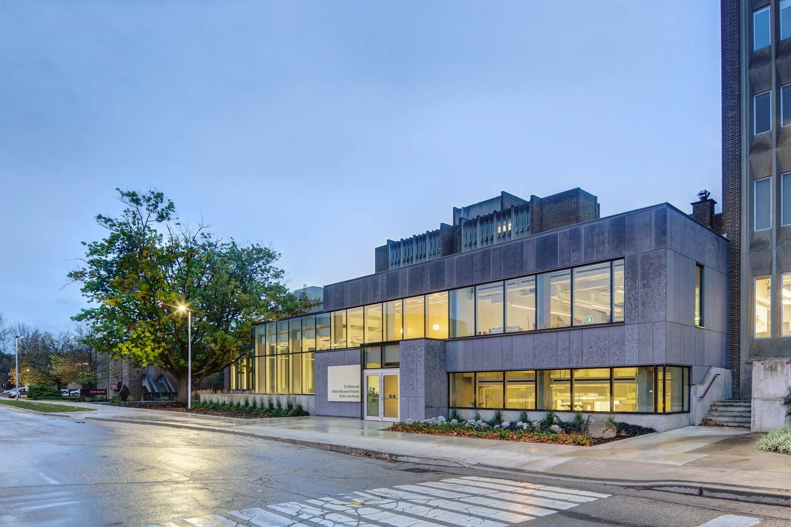 A modern building with large glass windows on a street during early evening, with a tree and streetlights nearby.