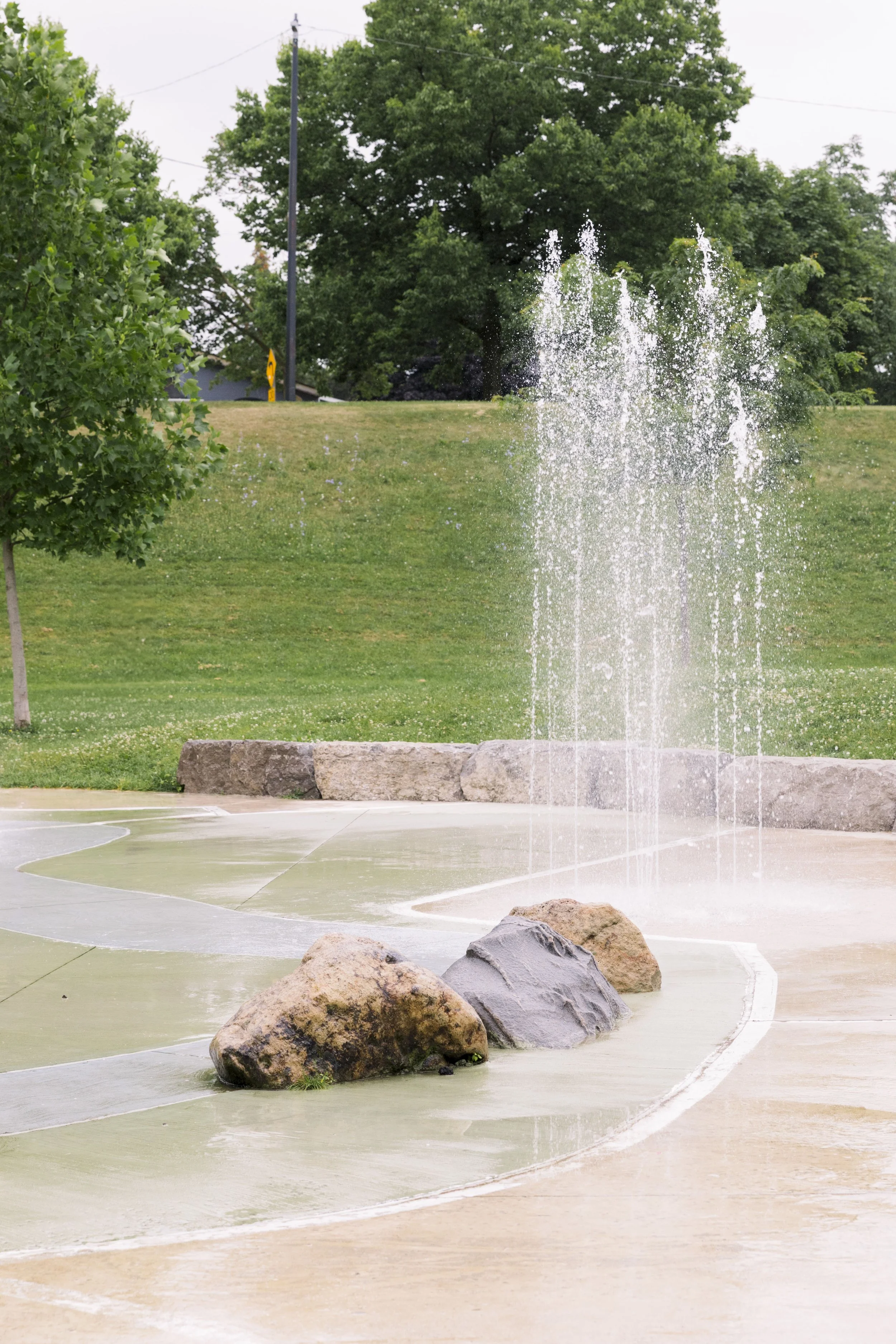A park with a fountain spraying water, surrounded by rocks and green grass, with trees and utility poles in the background.