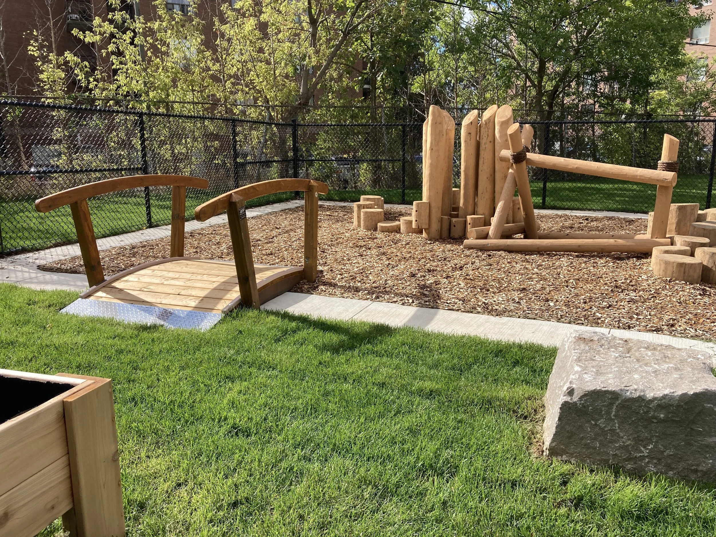 A playground with wooden structures, including a small bridge, logs, and a balancing beam, surrounded by green grass, a wood chip ground cover, a black chain-link fence, trees, and buildings in the background.
