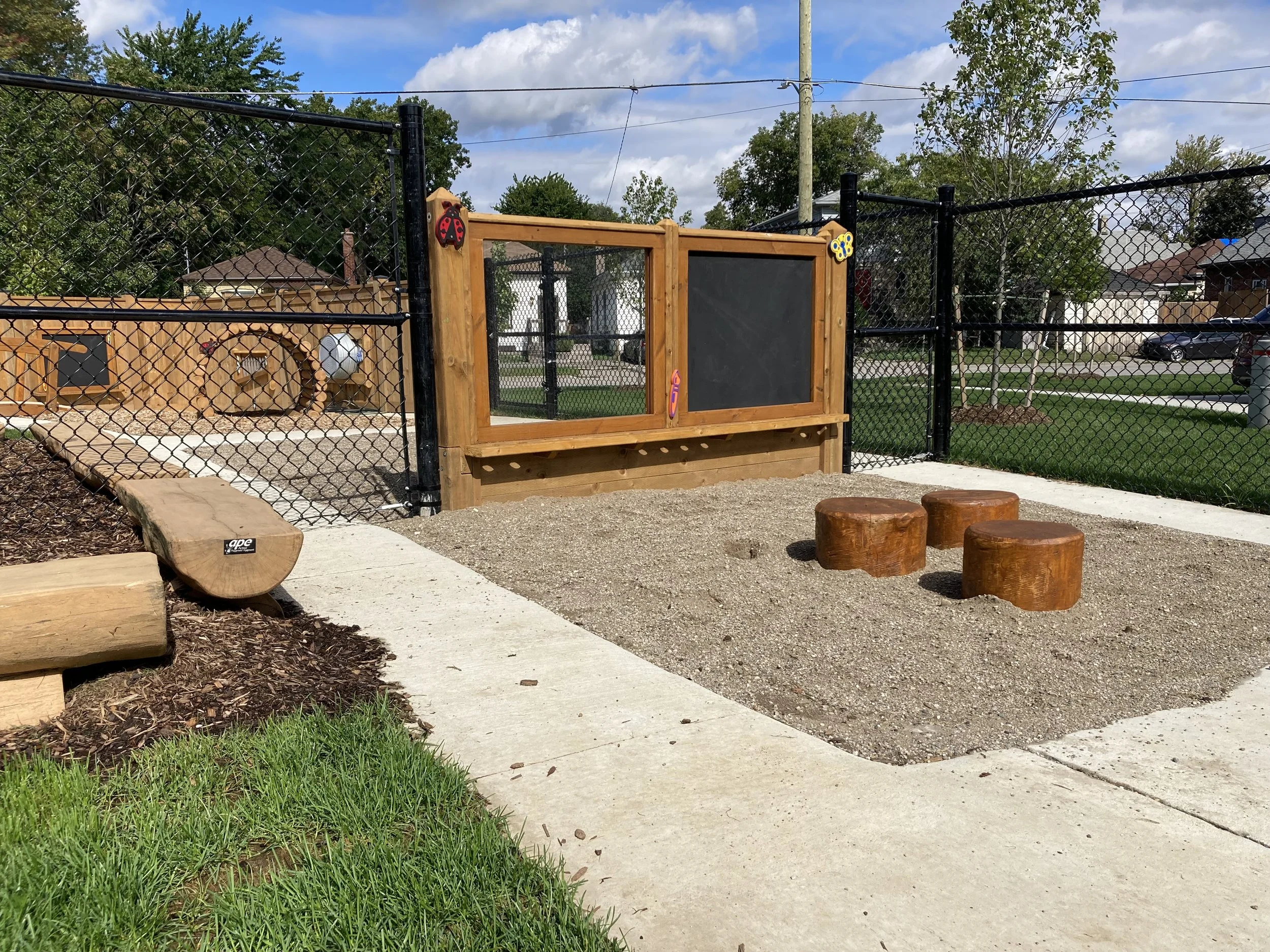 An outdoor playground area with a black chain-link fence, three wooden stepping stools, a wooden structure with a chalkboard, a rock climbing wall, and a small wooden house in the background. The area is surrounded by trees, grass, and nearby residen