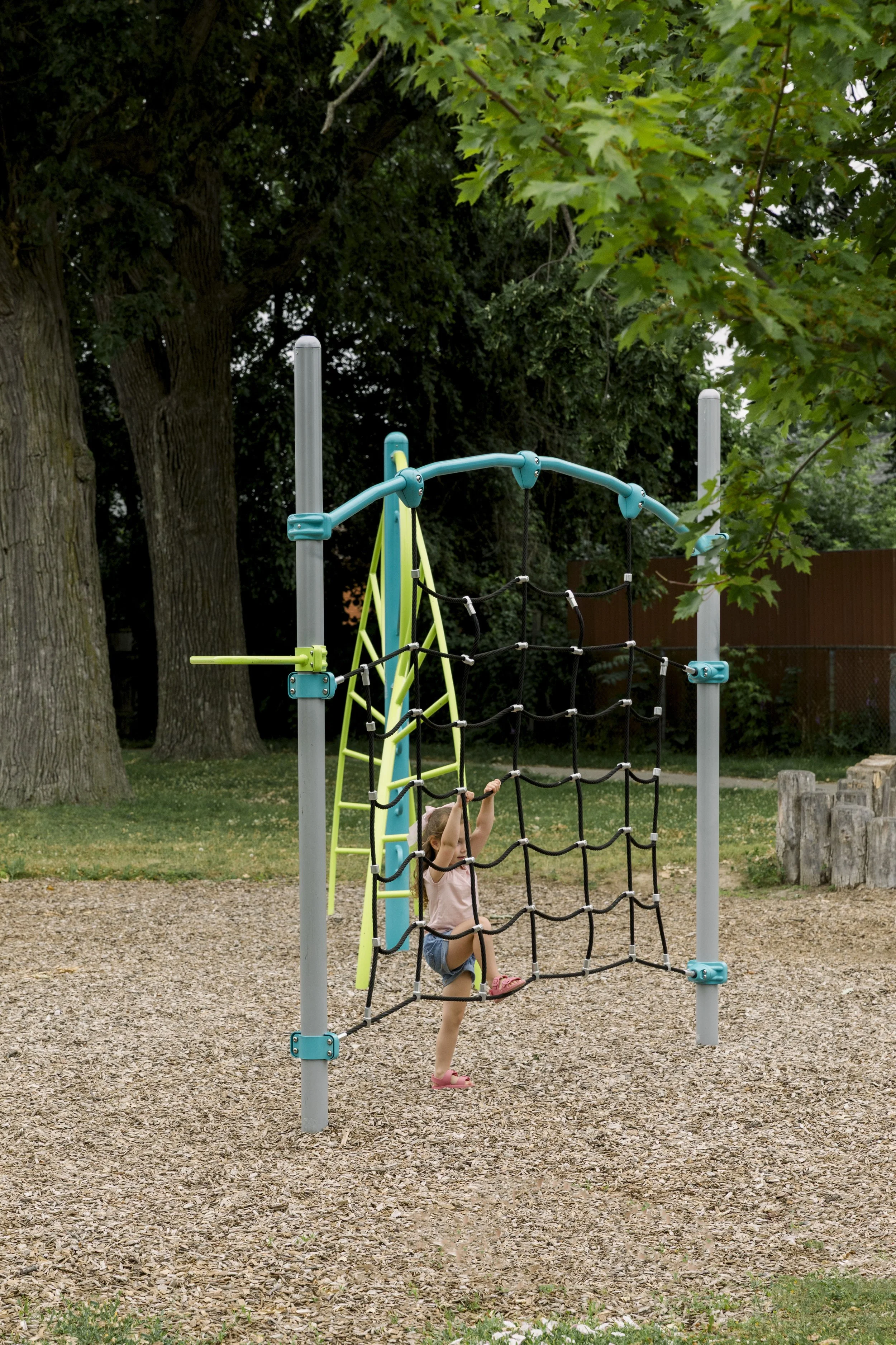Young girl climbing on a black rope ladder of a playground structure in a park with trees in the background.