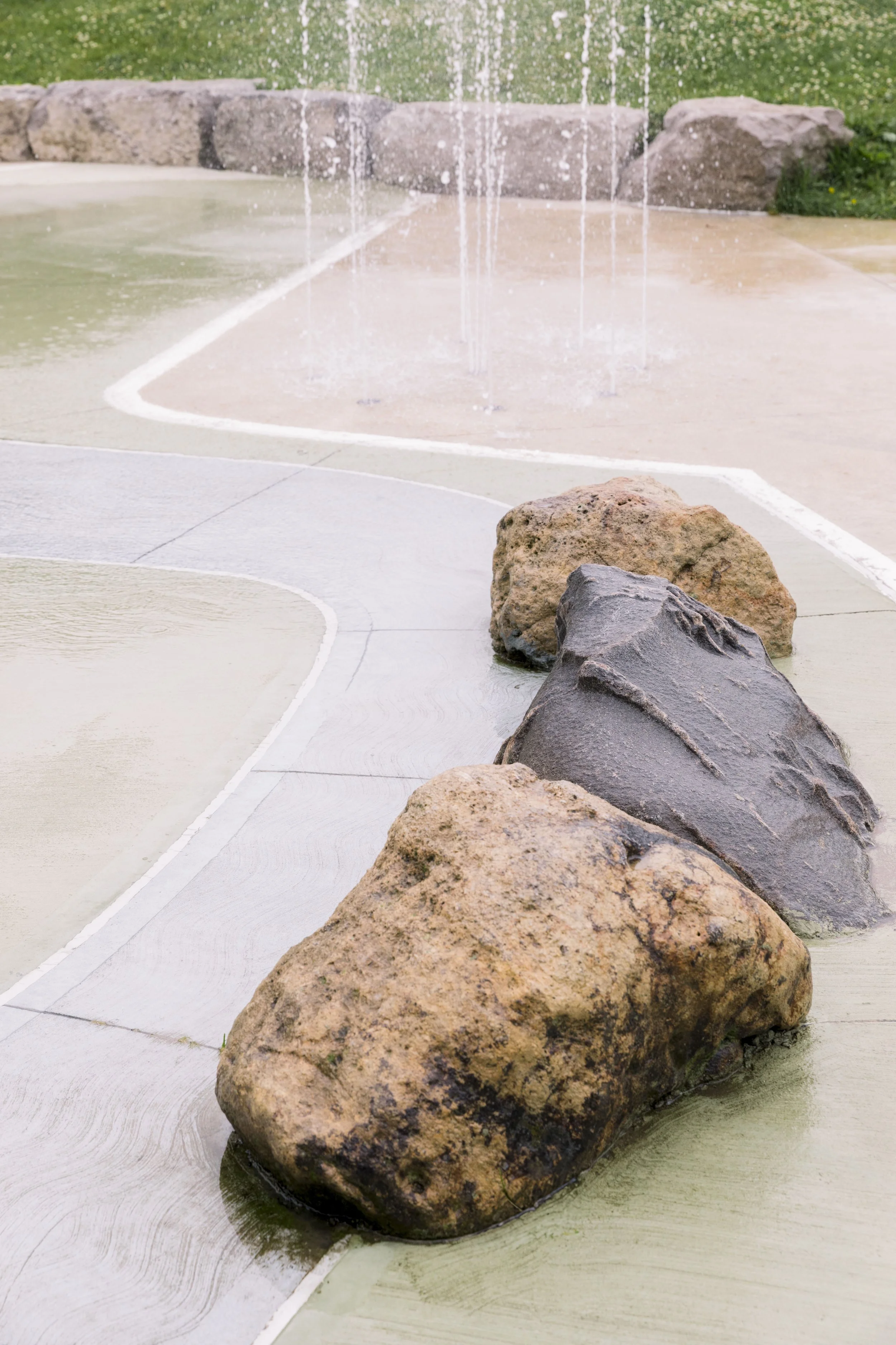 Boulders surrounded by decorative concrete with water fountain in background at park.
