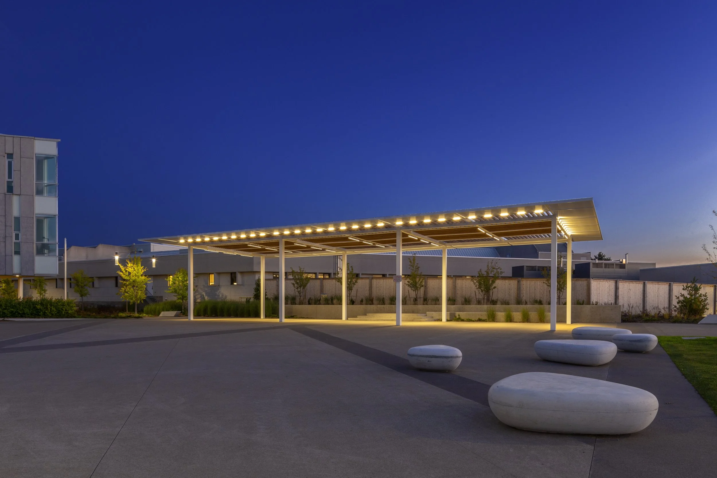 Modern outdoor pavilion with white supporting columns and a flat roof illuminated with strip lighting, surrounded by small trees and stylized white stone seating, during twilight with an urban building in the background.