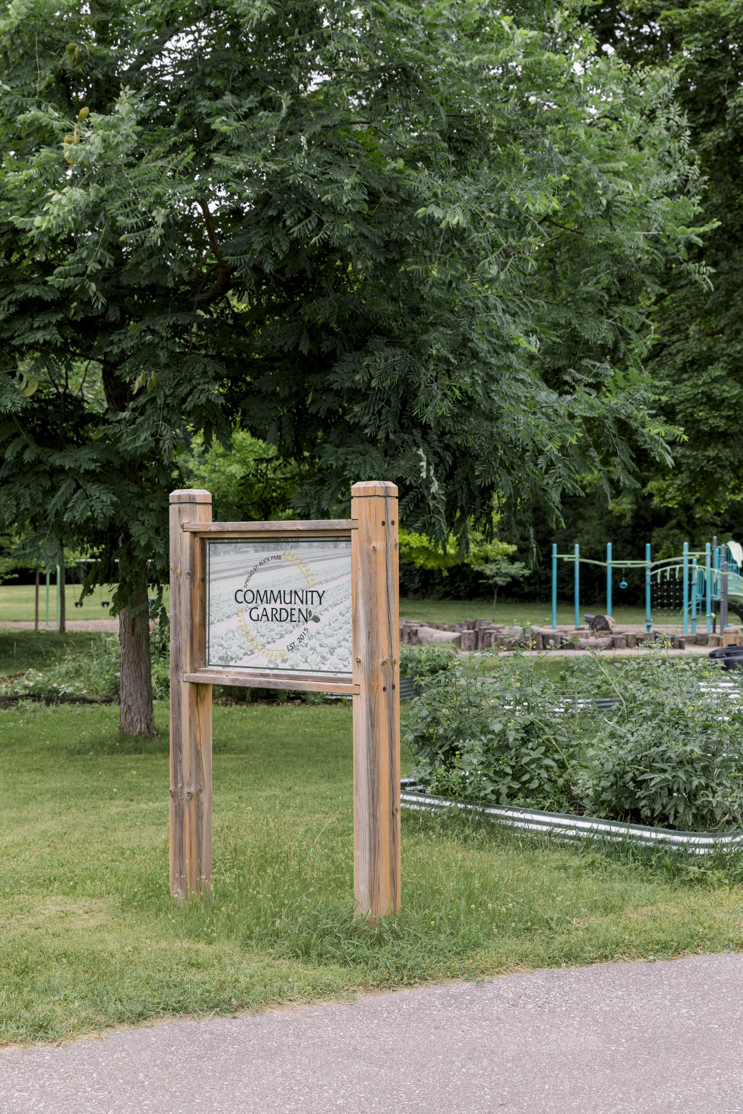 A sign for a community garden with a tree and playground in the background.