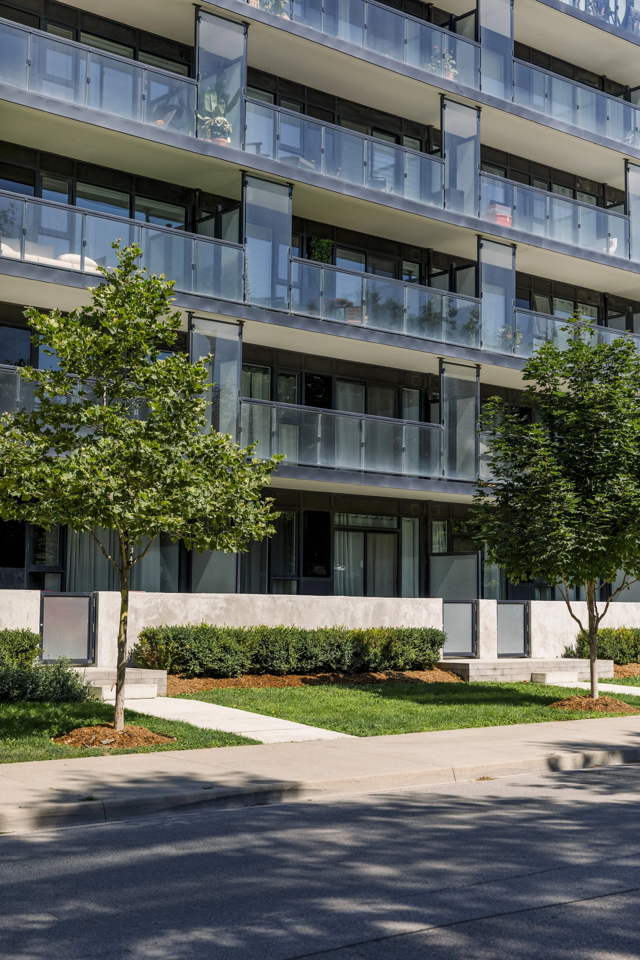 Modern apartment building with glass balconies, trees, and a sidewalk.