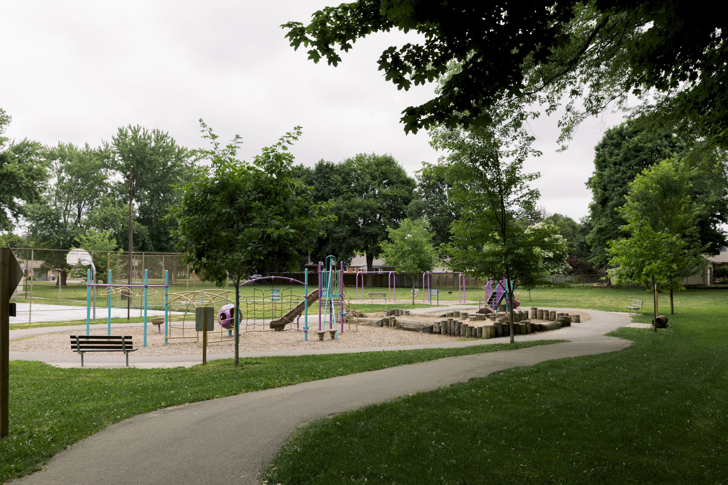 Empty playground in a park with trees, a paved path, benches, and a fenced basketball court on a cloudy day.