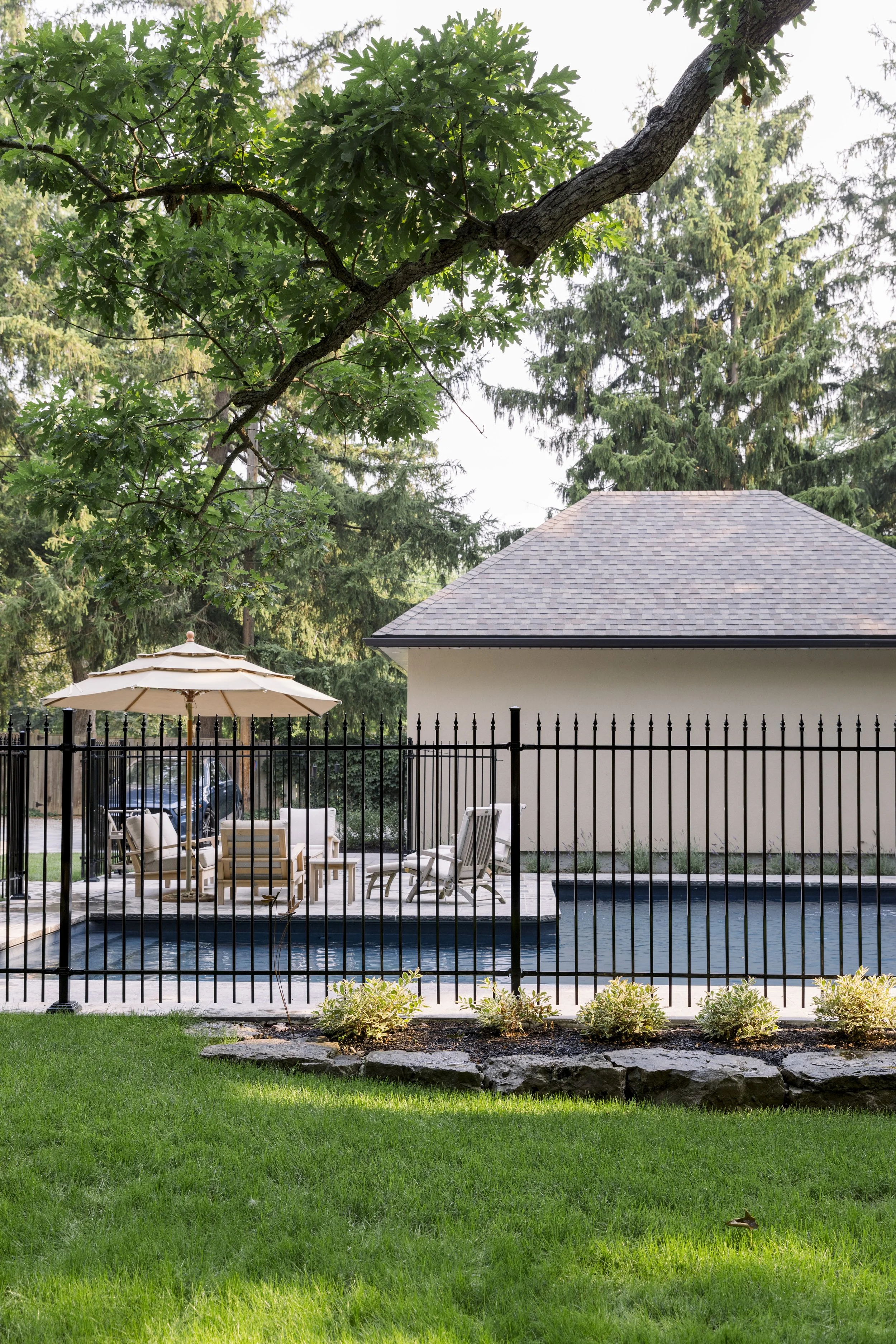Backyard scene with a swimming pool surrounded by a black metal fence, patio furniture including chairs and a table with an umbrella, and a small beige building. Green grass and trees in the background.