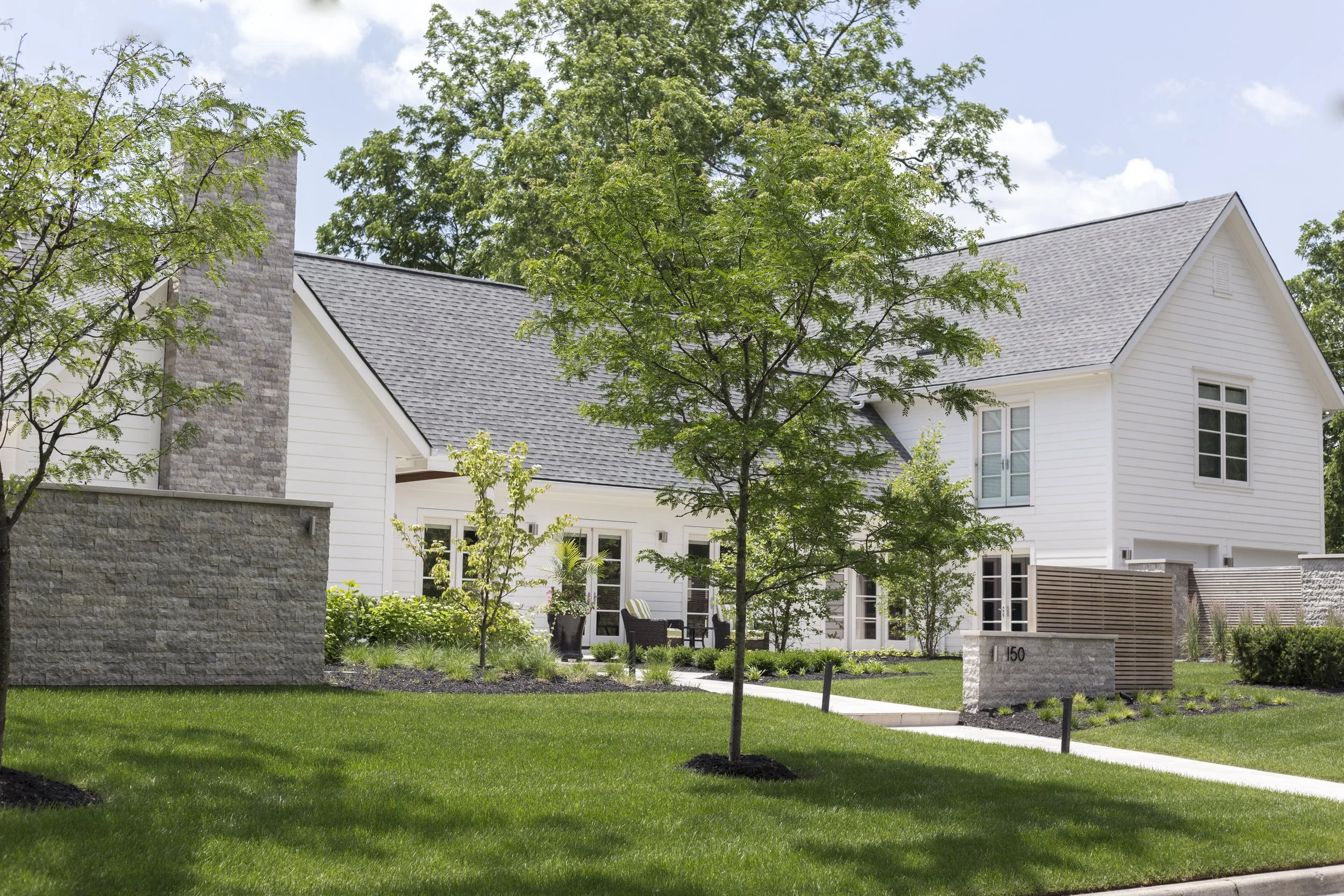 A modern white house with multiple windows, surrounded by a manicured lawn and young trees, with a sidewalk in front and partly cloudy sky overhead.