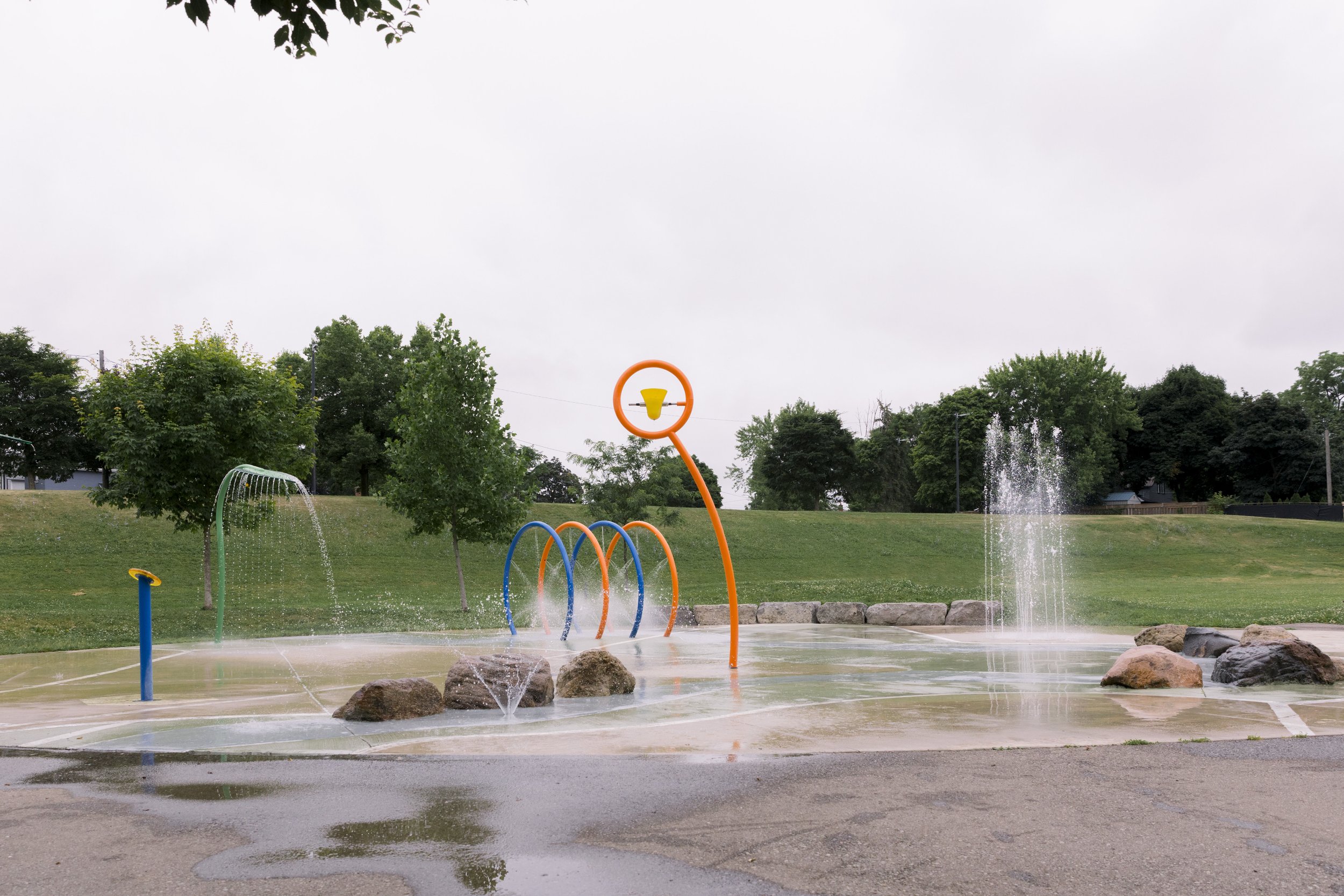 Empty splash pad with water features, including sprayers, fountains, and a circular colorful arch, on a cloudy day with wet pavement and green trees in the background.