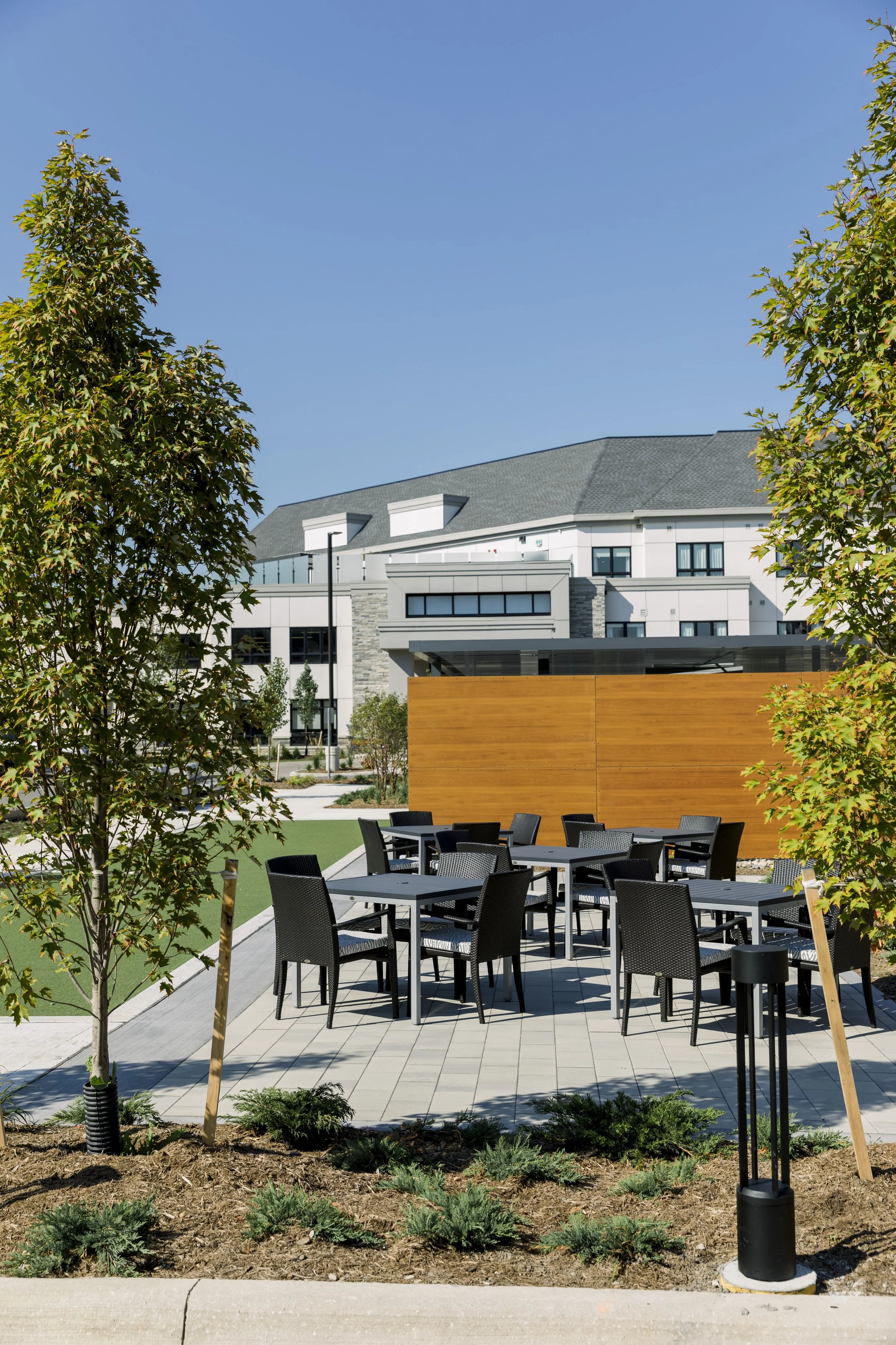 Outdoor patio area with black chairs and tables, surrounded by trees and a wooden privacy screen, with a modern multi-story building in the background under a clear blue sky.