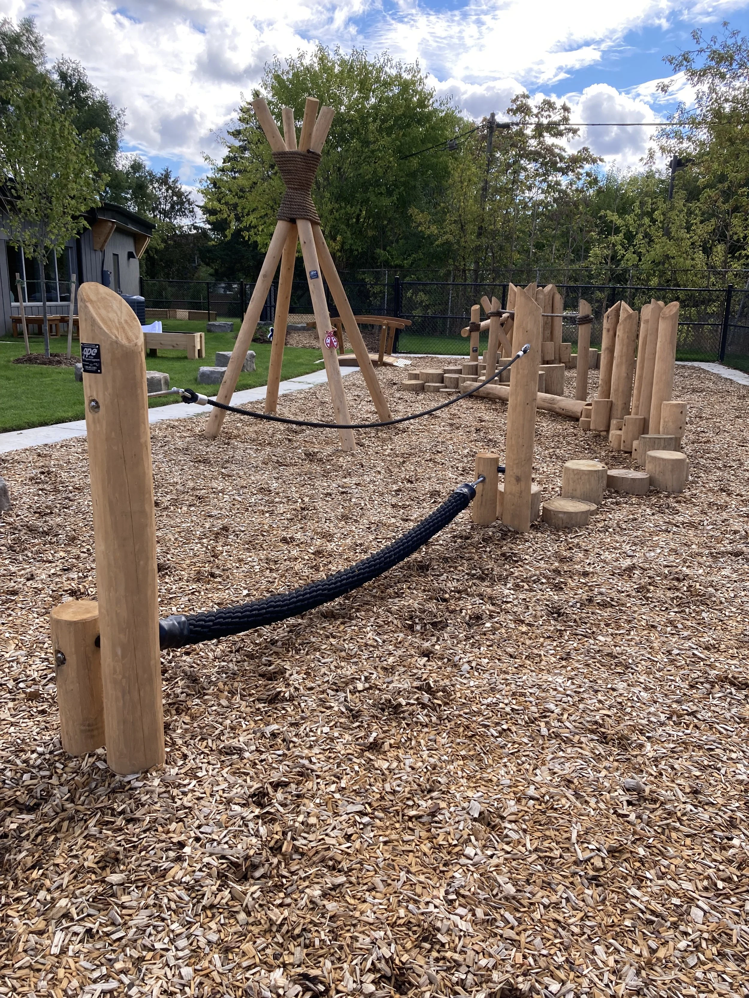 Wooden playground with a climbing wall, logs, and a tipi-style structure surrounded by wood chips, with a grassy area and trees in the background.