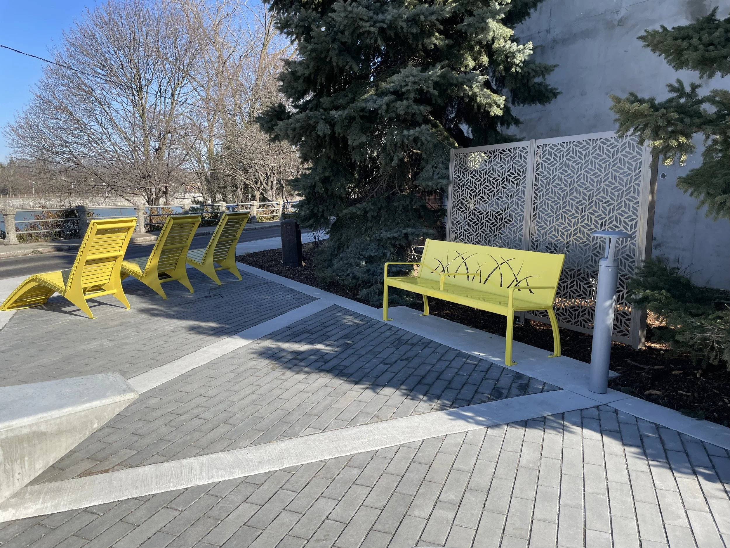 Four yellow lounge chairs and a yellow bench with a decorative metal screen behind, in a sunny outdoor area with trees and a sidewalk.