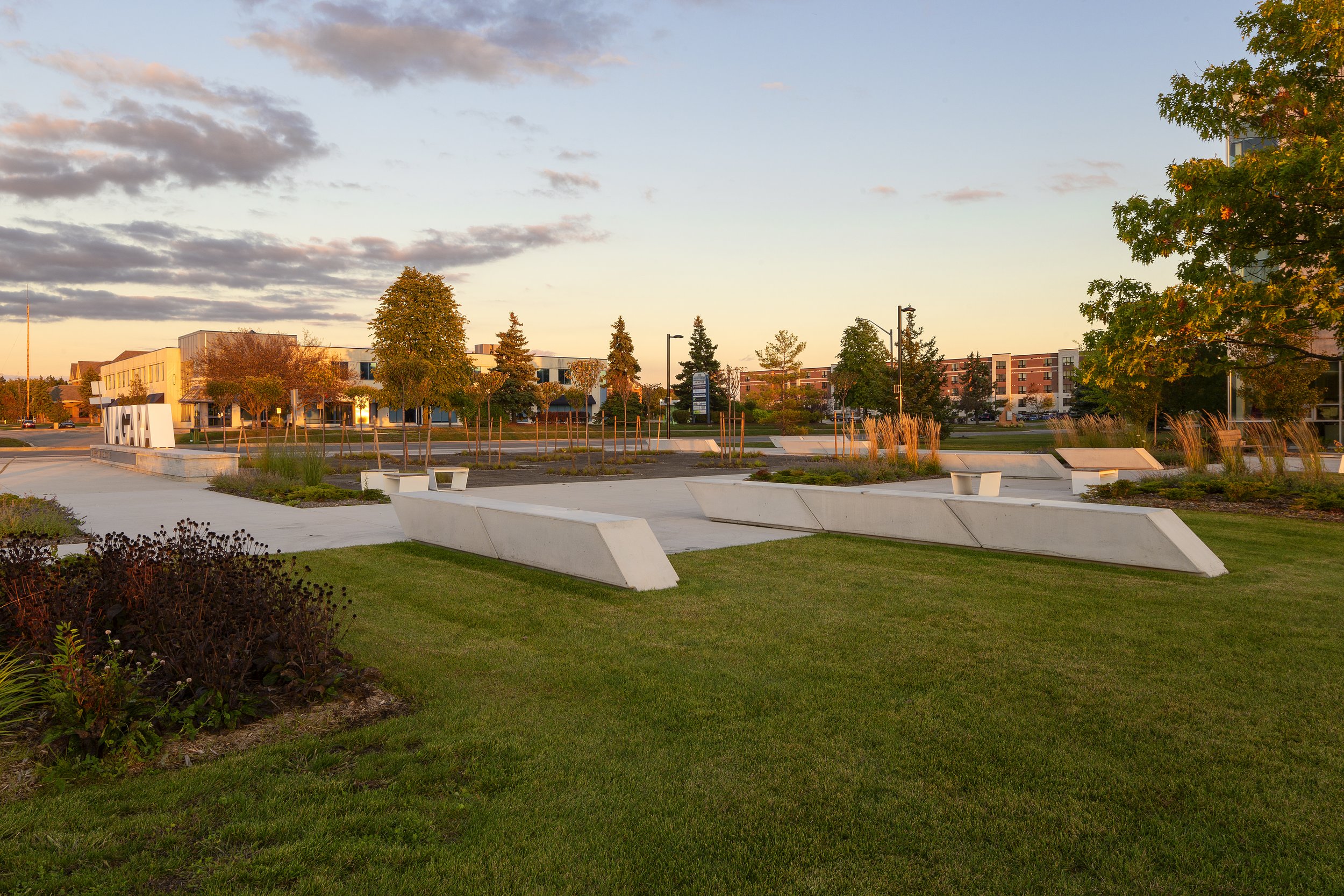 A park with a modern sculpture displaying the word 'UMCH' in large white letters, concrete benches, and green landscaping under a partly cloudy sky at sunset.