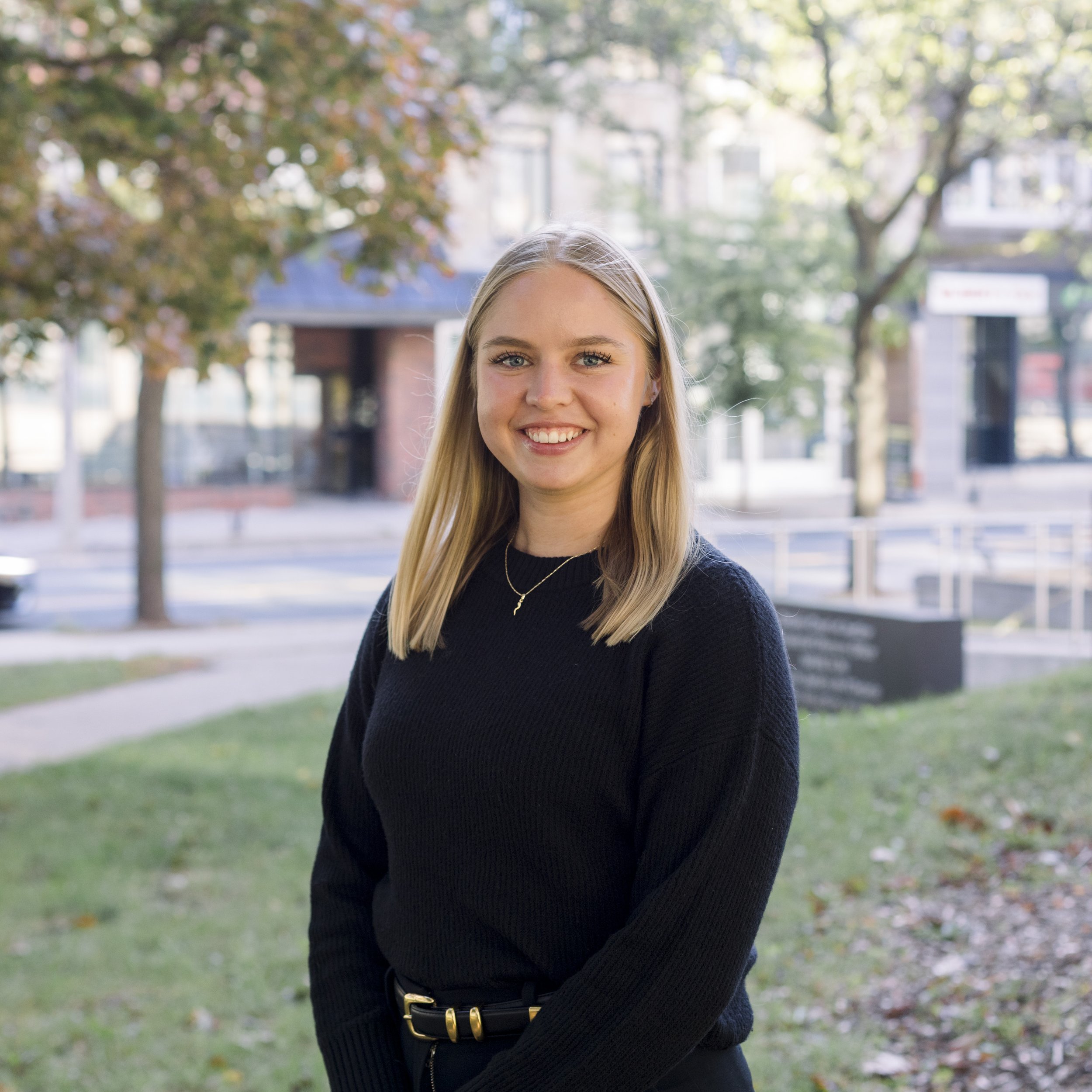 A smiling young woman with blonde hair in a black sweater standing outdoors in a park with trees and buildings in the background.
