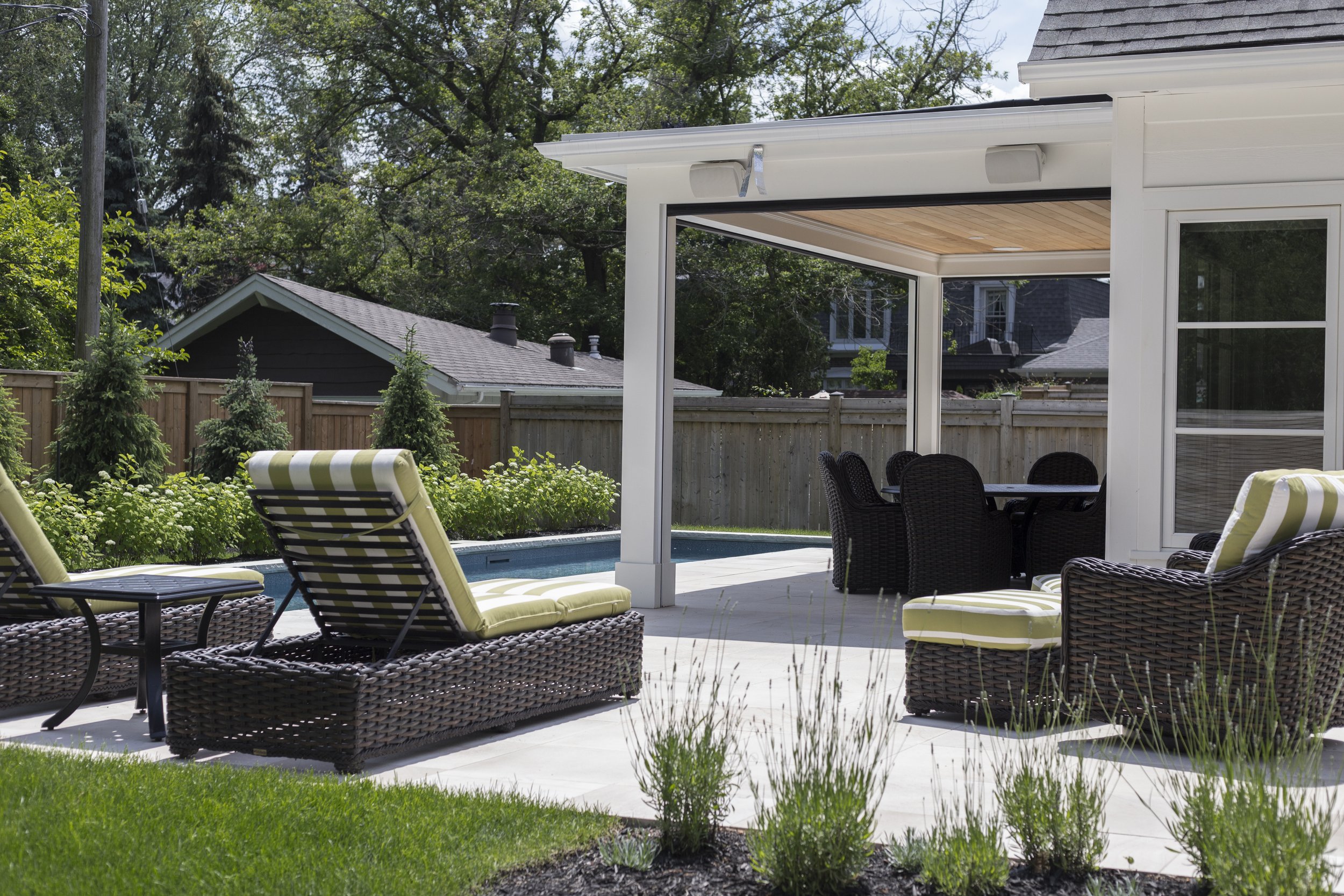 Backyard patio with lounge chairs, a table, a dining table with chairs under a covered area, a swimming pool, and a landscaped yard with trees and shrubs.