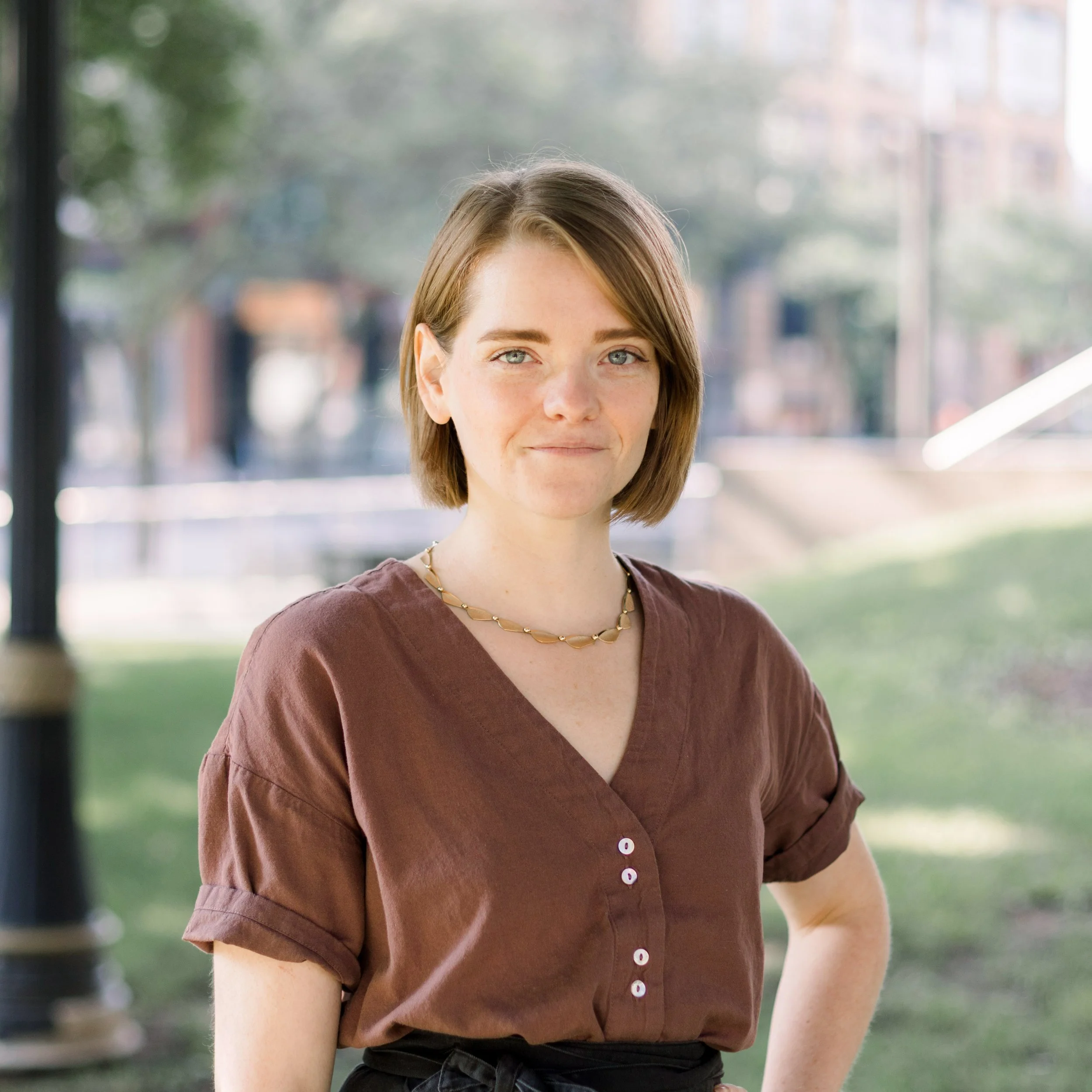 A young woman with short brown hair and blue eyes standing outdoors near a glass building, wearing a brown short-sleeved blouse and a gold necklace.