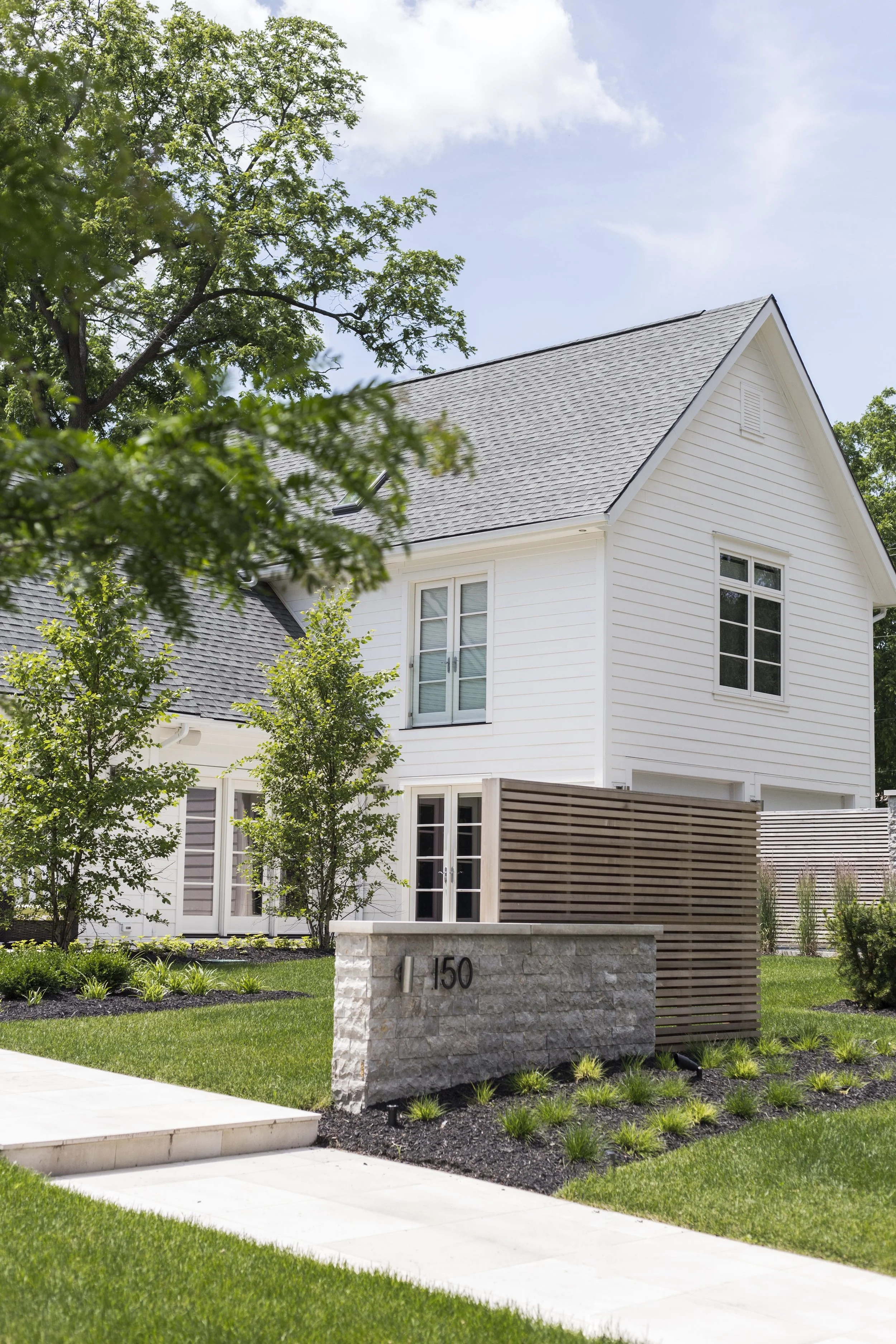 Front view of a modern two-story house with white siding, large windows, and a gray shingled roof, surrounded by a neatly manicured lawn and trees, with a sidewalk and a stone mailbox.