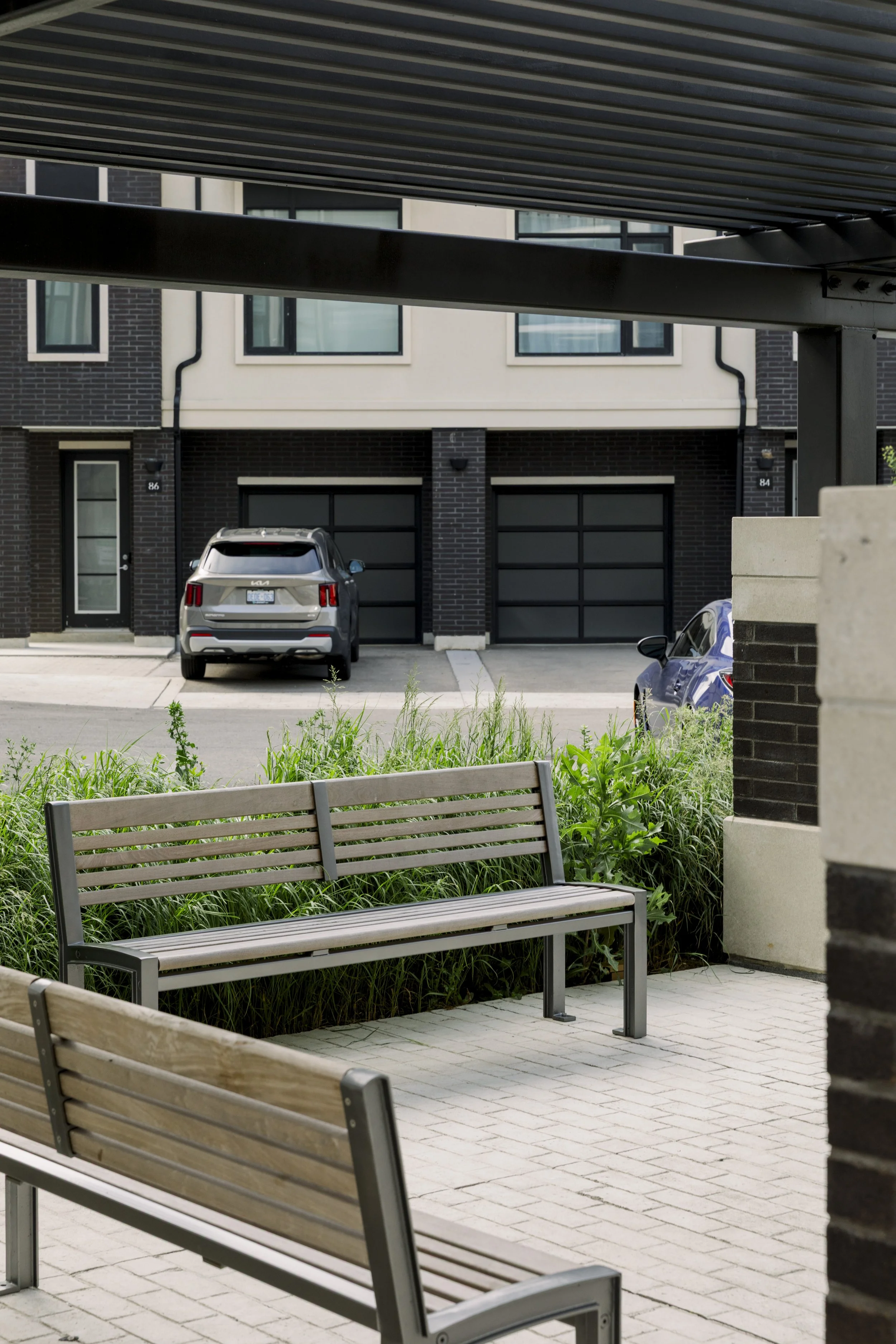 Residential parking lot with two parked cars, modern apartment buildings in the background, and wooden benches with surrounding greenery in the foreground.