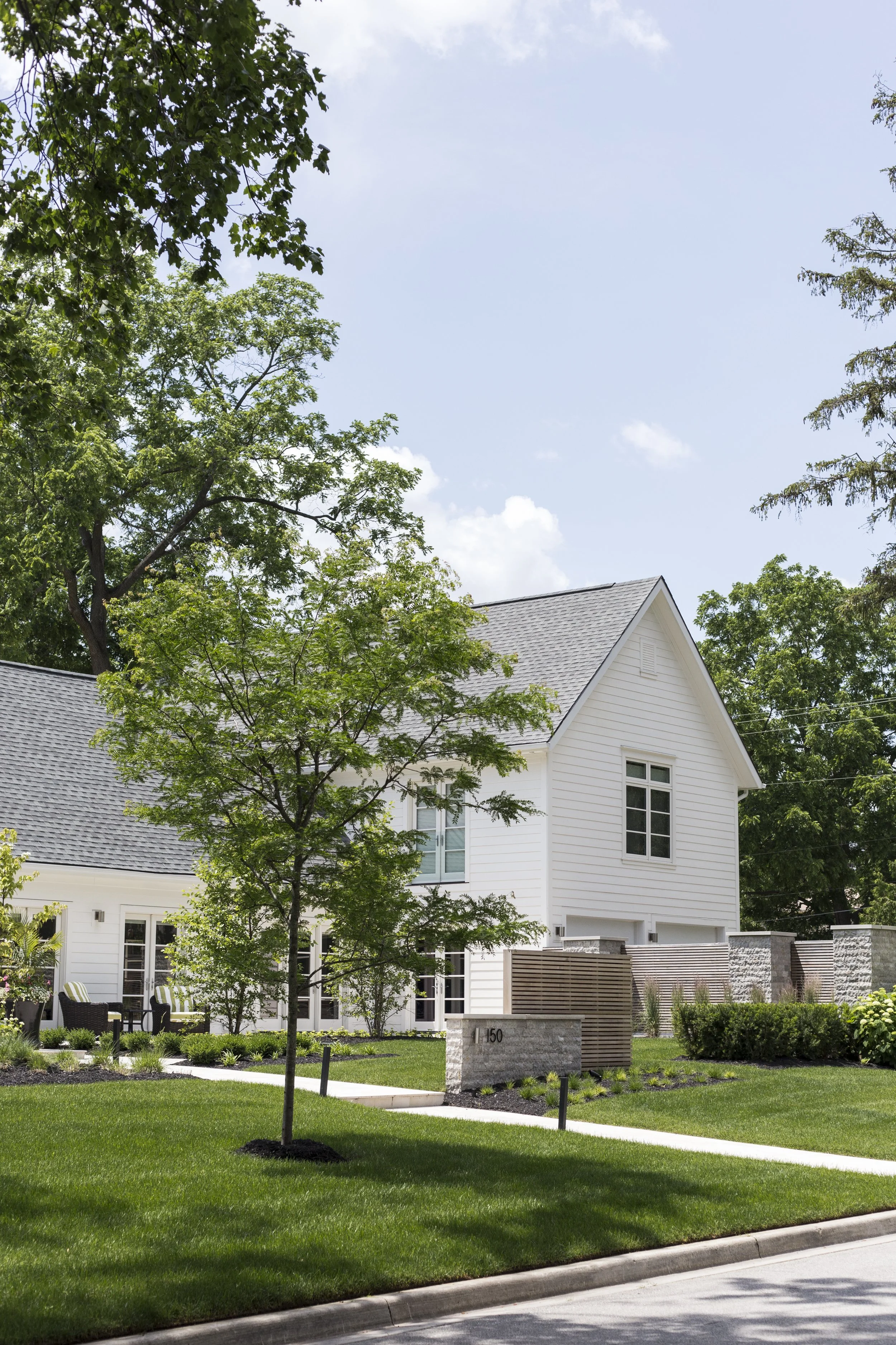 Modern white house with a gray roof, surrounded by green trees and grass, on a sunny day.