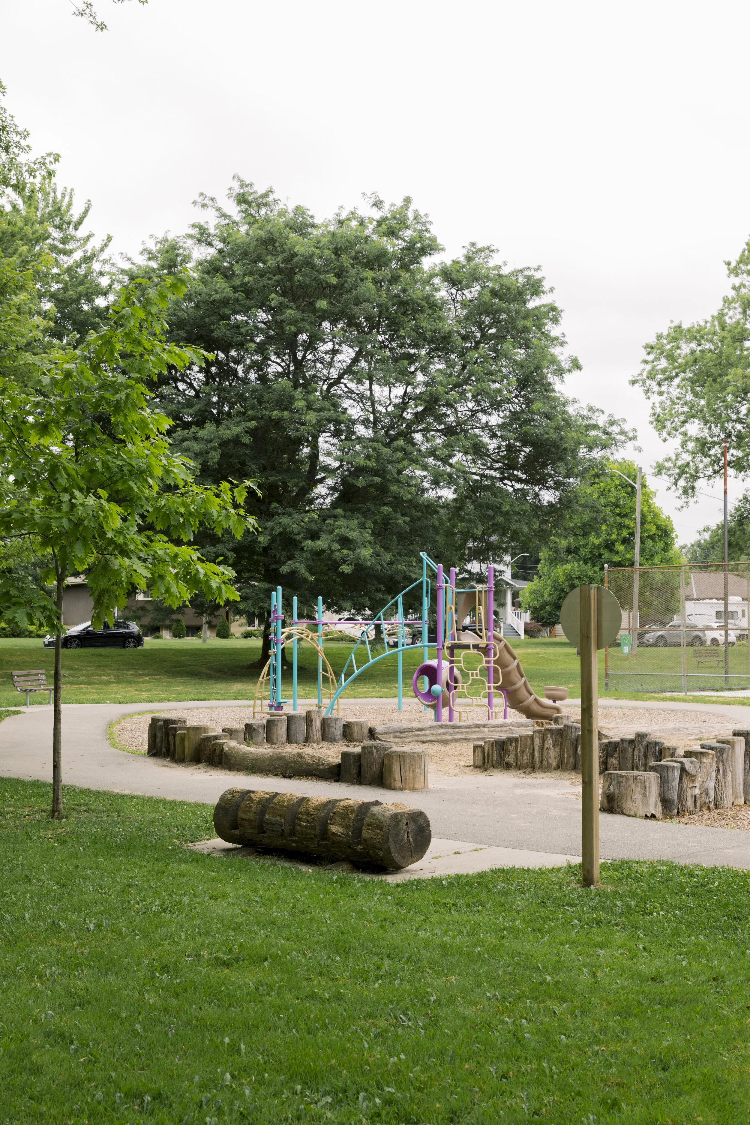 Empty playground with jungle gym, slide, and wooden logs in a park with green grass and trees on overcast day.