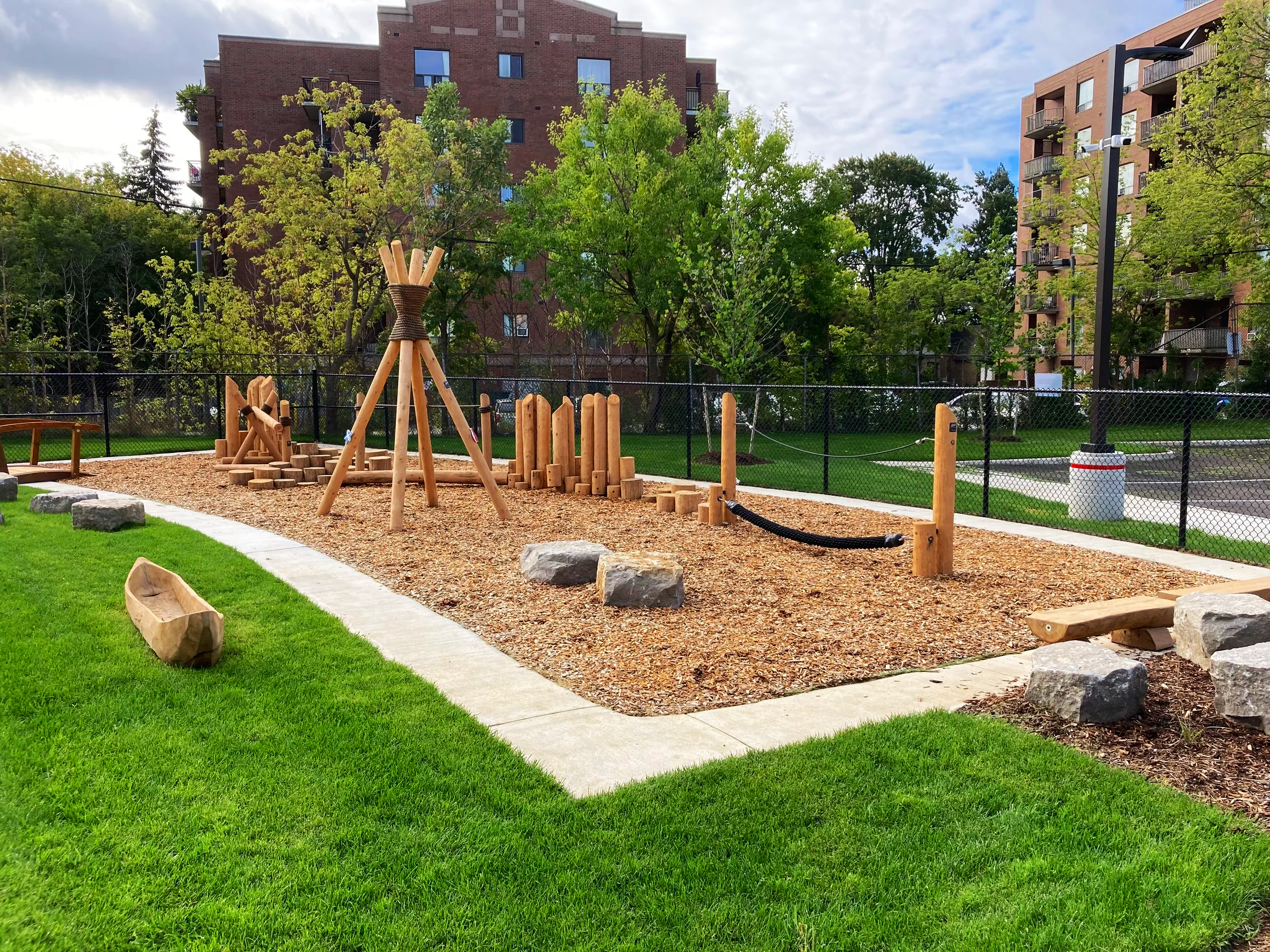 Children's playground with wooden structures, rocks, and a rope bridge, surrounded by a grassy area and residential apartment buildings in the background.
