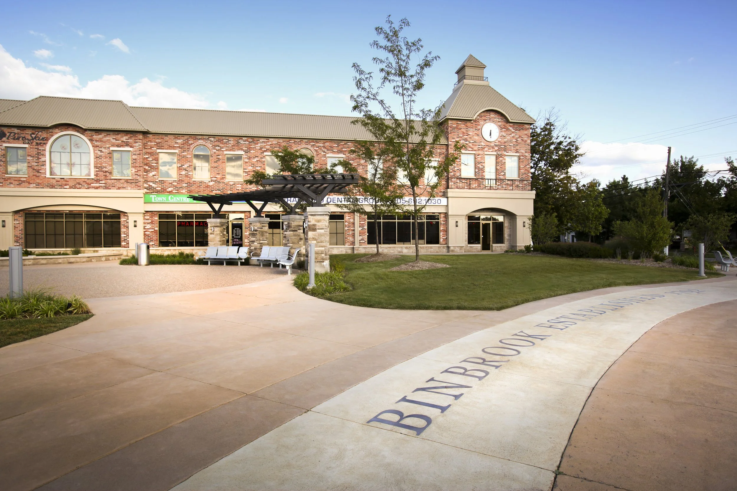 A shopping center with a brick facade, arched windows, and a clock tower, surrounded by a landscaped area with trees, benches, and a walkway with inscribed text.