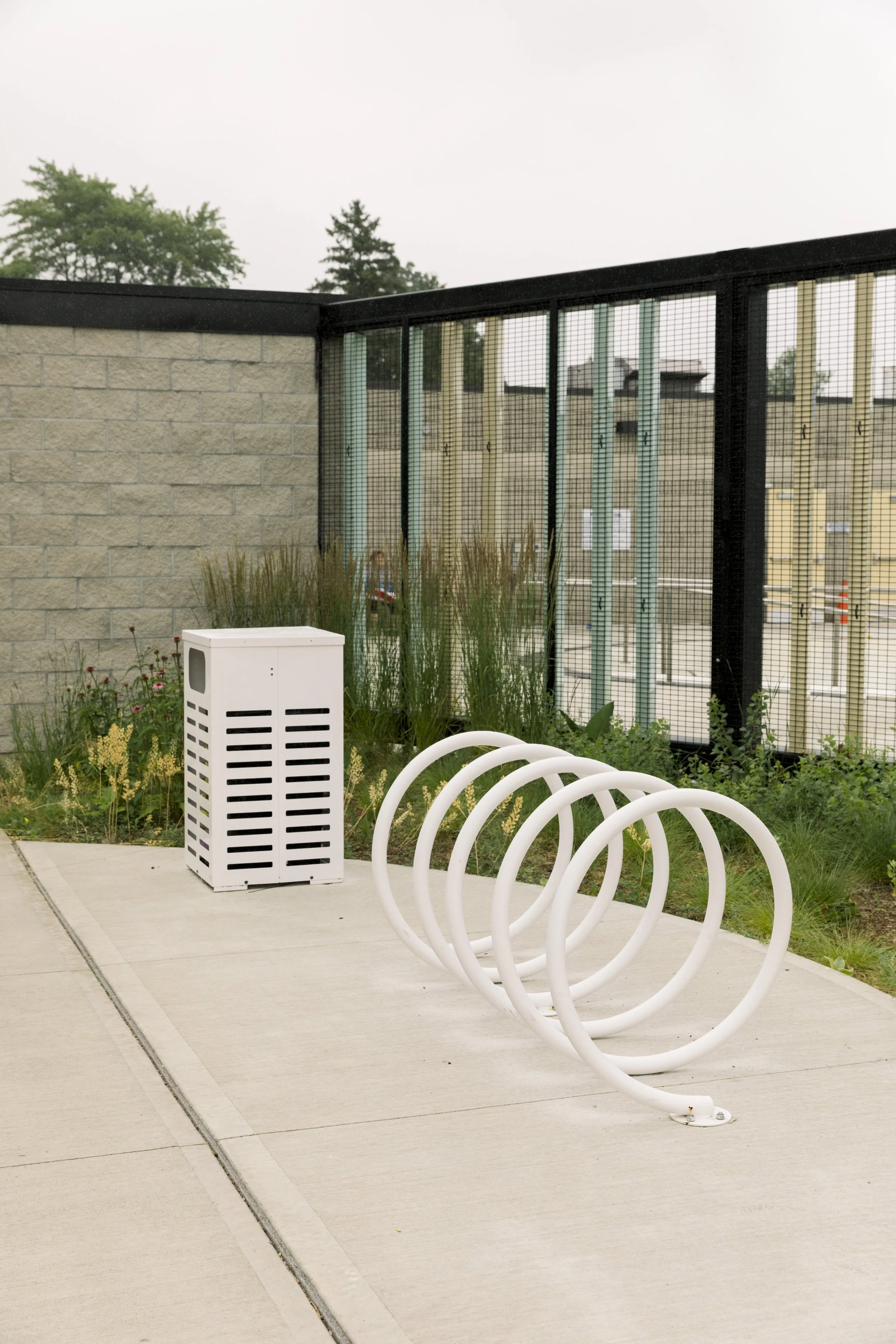 Bike rack, air conditioning unit, and plants on sidewalk near a metal fence and brick wall.