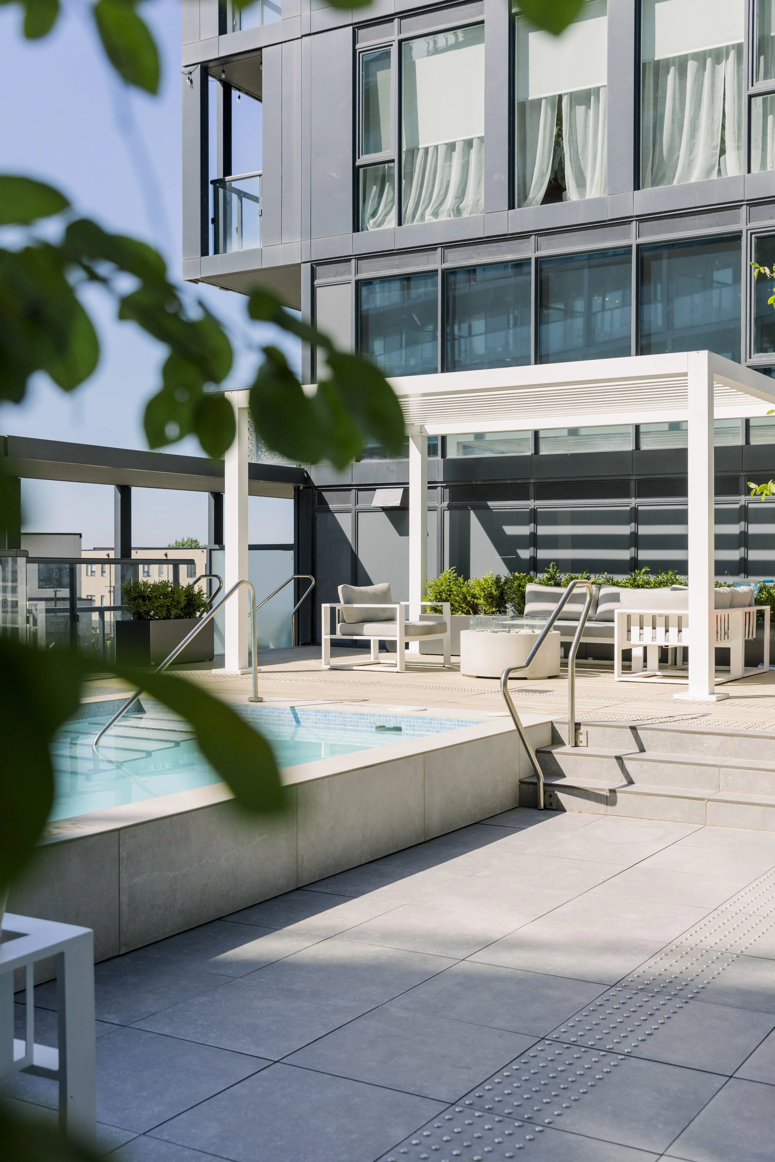 Outdoor rooftop pool area with modern patio furniture, surrounded by glass buildings and greenery, under a clear blue sky.
