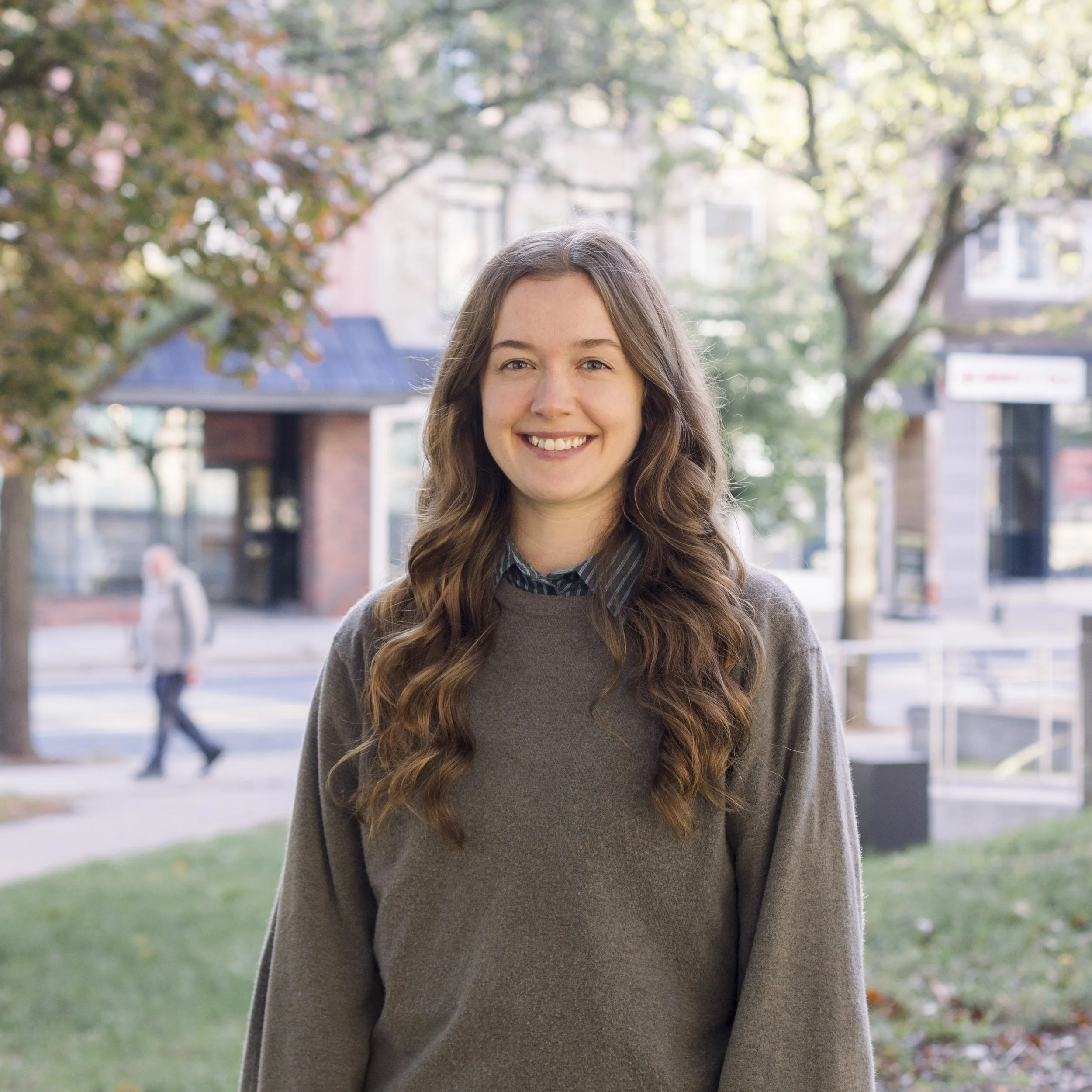 A young woman with long, wavy brown hair smiling outdoors on a sidewalk, with trees and buildings in the background.