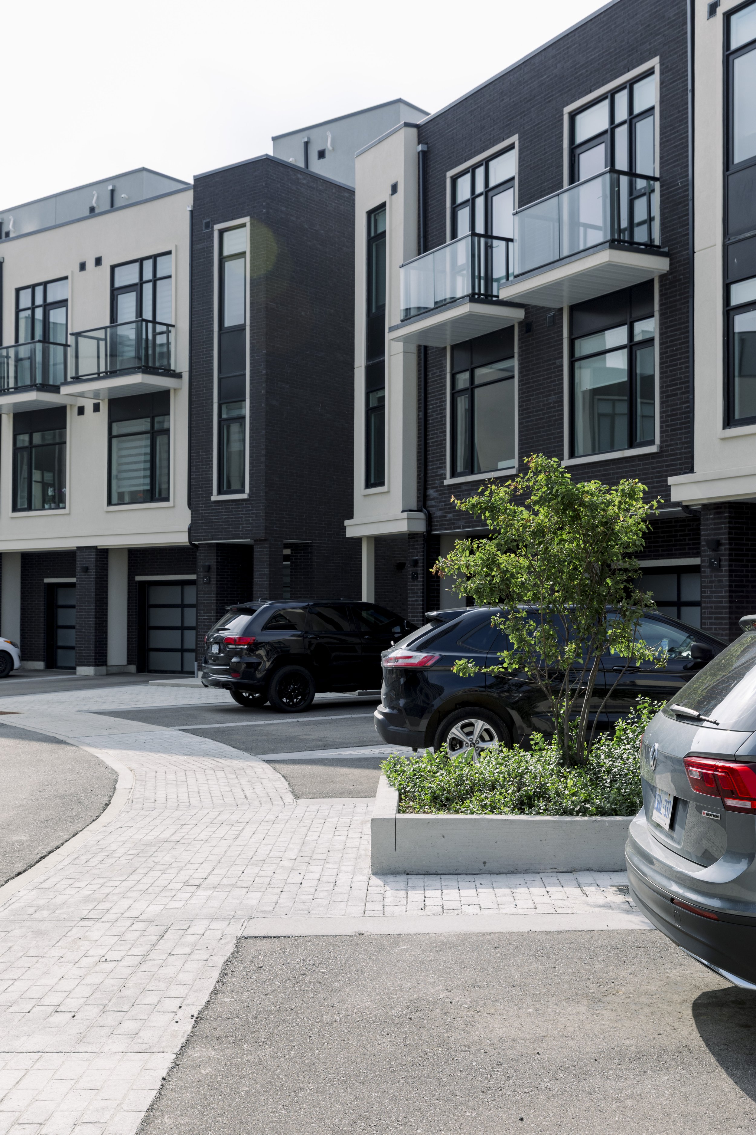 Modern apartment buildings with balconies and parked cars in front, small landscaped trees and sidewalks in a contemporary urban setting.