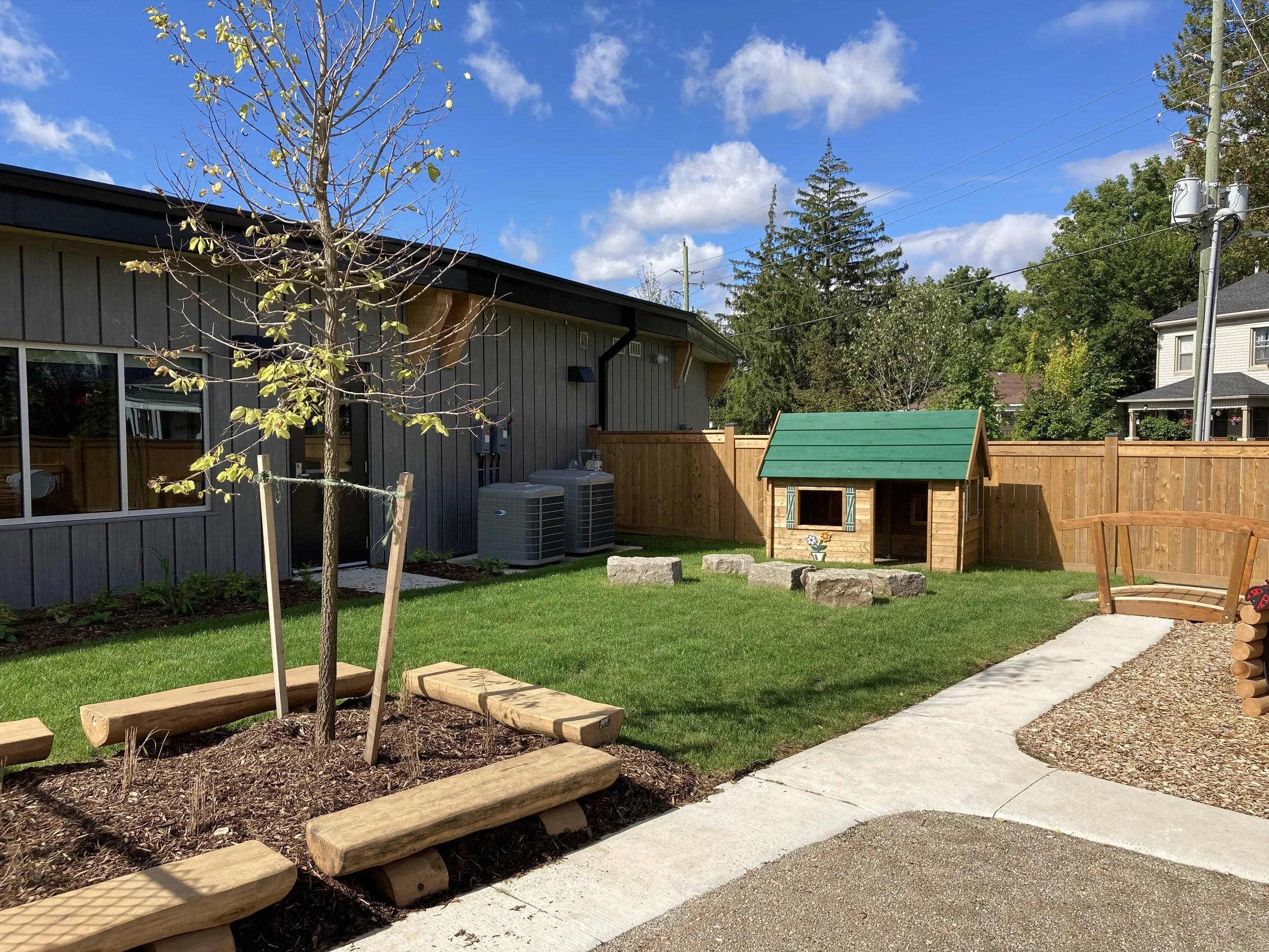 Backyard with a small tree, wood benches, a doghouse with a green roof, a wooden fence, and a paved walkway under a partly cloudy blue sky.