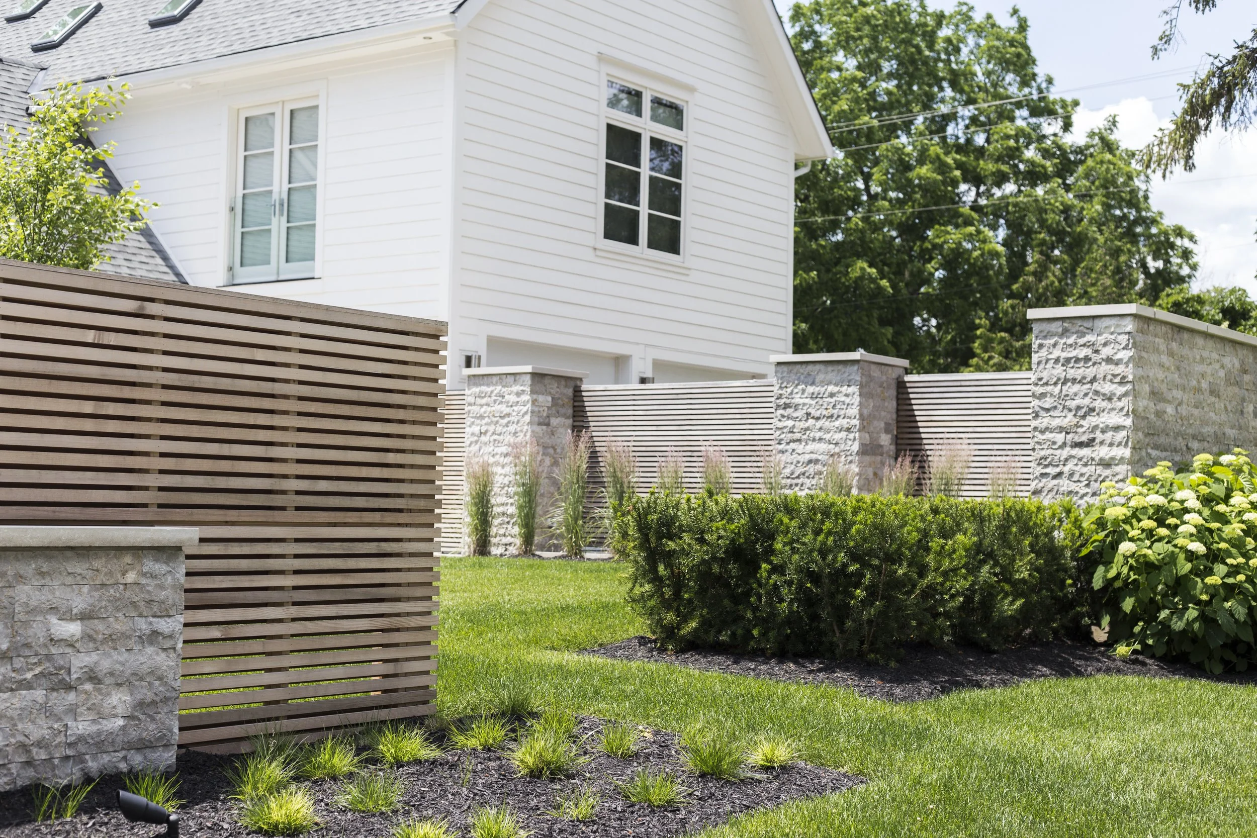 A modern white house with large windows, surrounded by a well-manicured lawn, shrubs, and a wooden and stone fence.