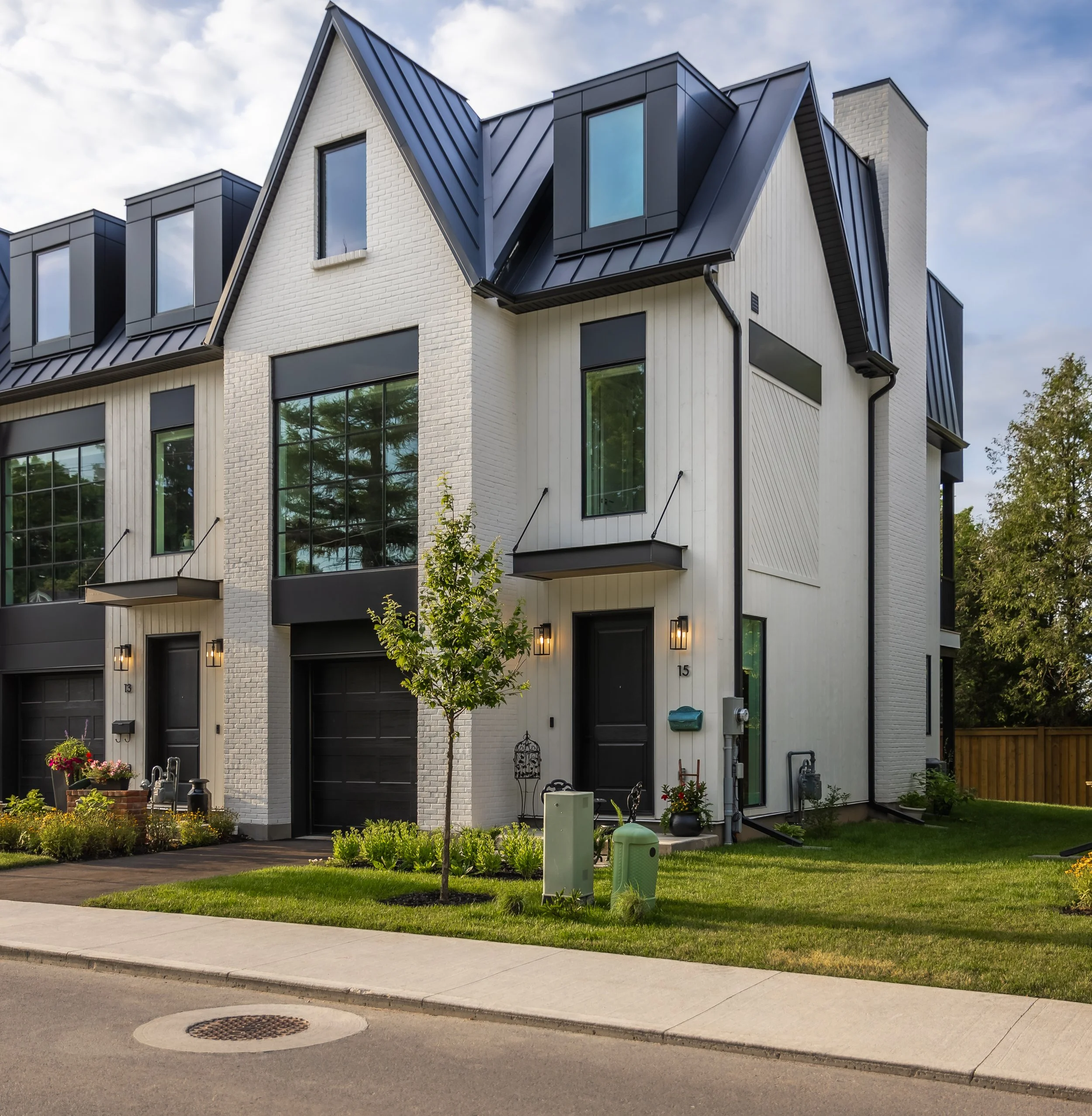 Modern multi-story townhouse with white brick and vertical siding, black accents, large windows, and a small front yard with a tree and landscaped plants