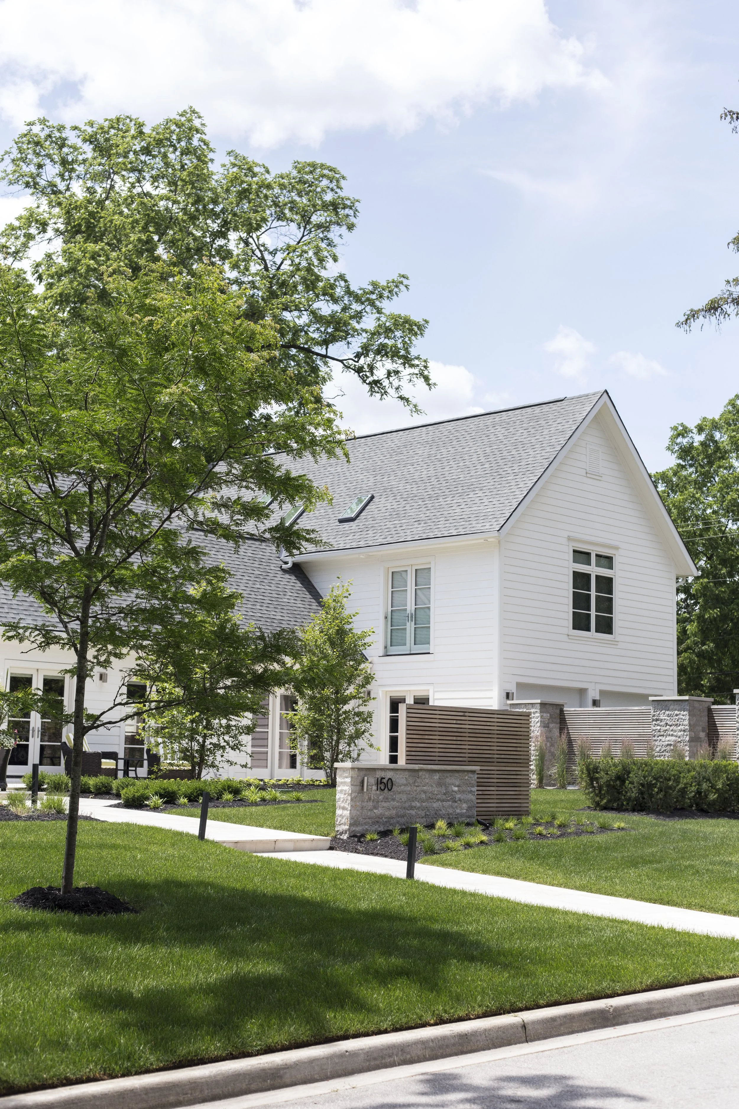 A modern white house with a gray roof, surrounded by a green lawn and trees, with a concrete sidewalk in the foreground.