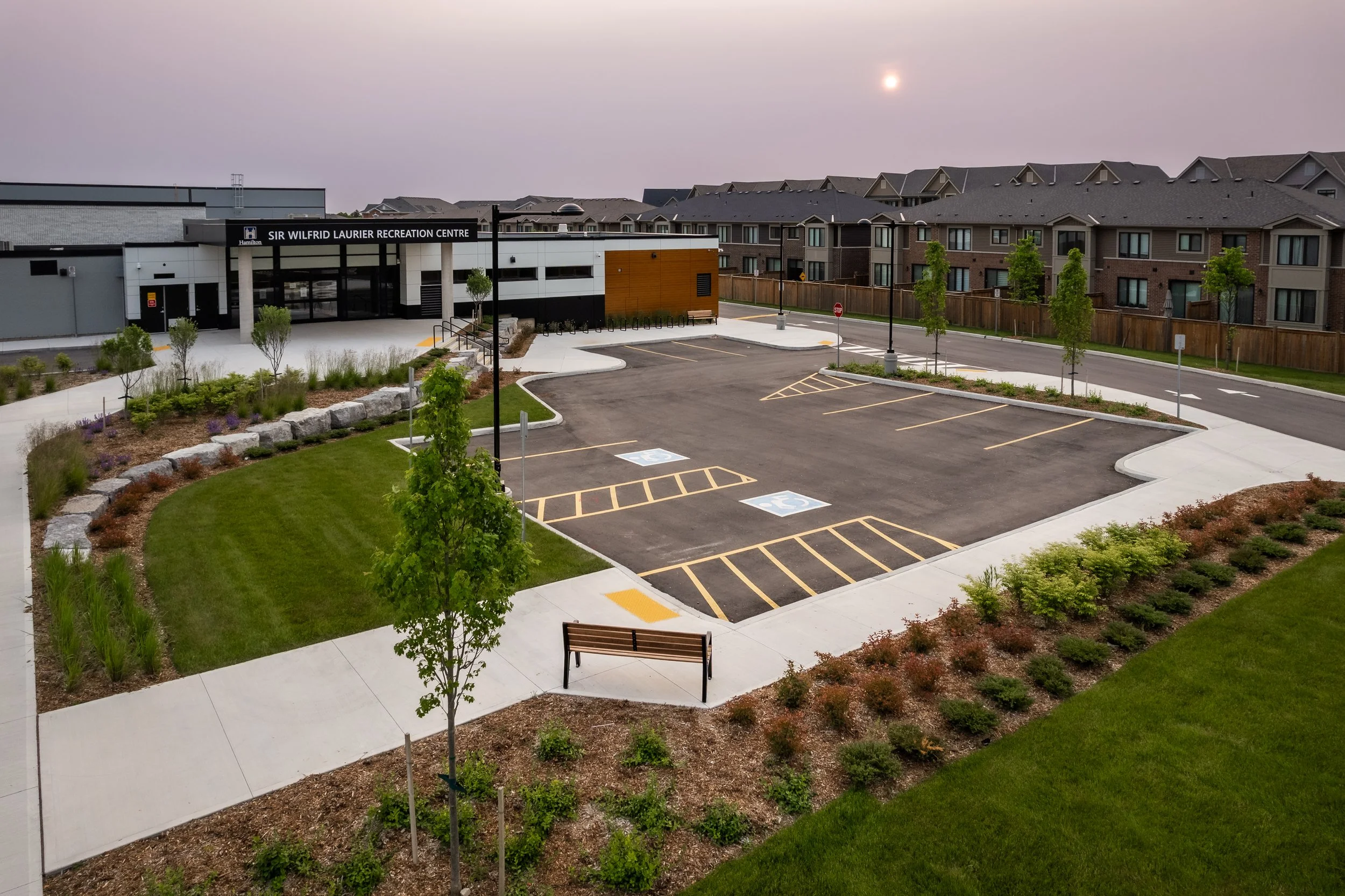 An empty parking lot with designated handicapped spaces, surrounded by landscaped grass and shrubs, with a building labeled Sir Wilfrid Laurier Recreation Centre in the background.