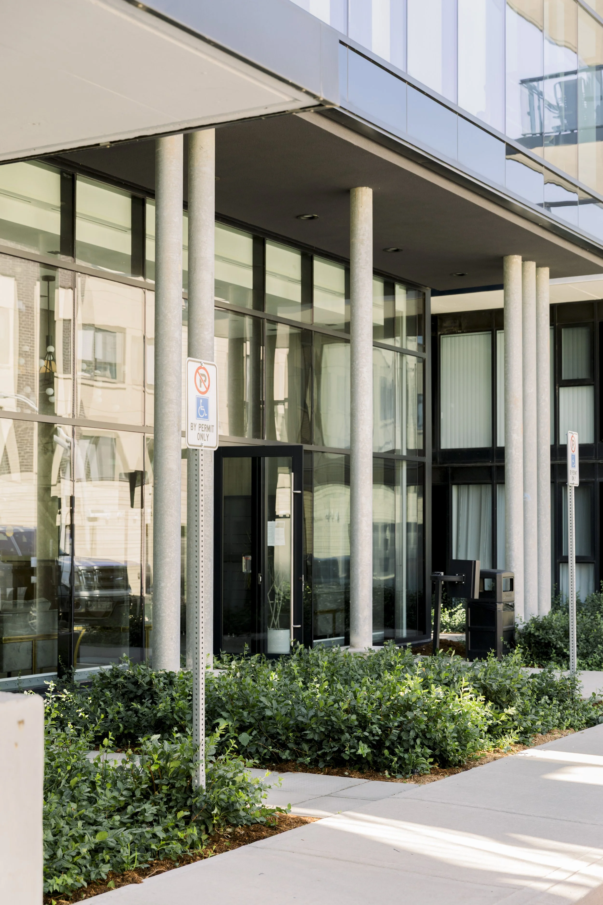 Modern glass-front building with metal columns, small shrubbery, sidewalk, and parking signs indicating handicapped parking.