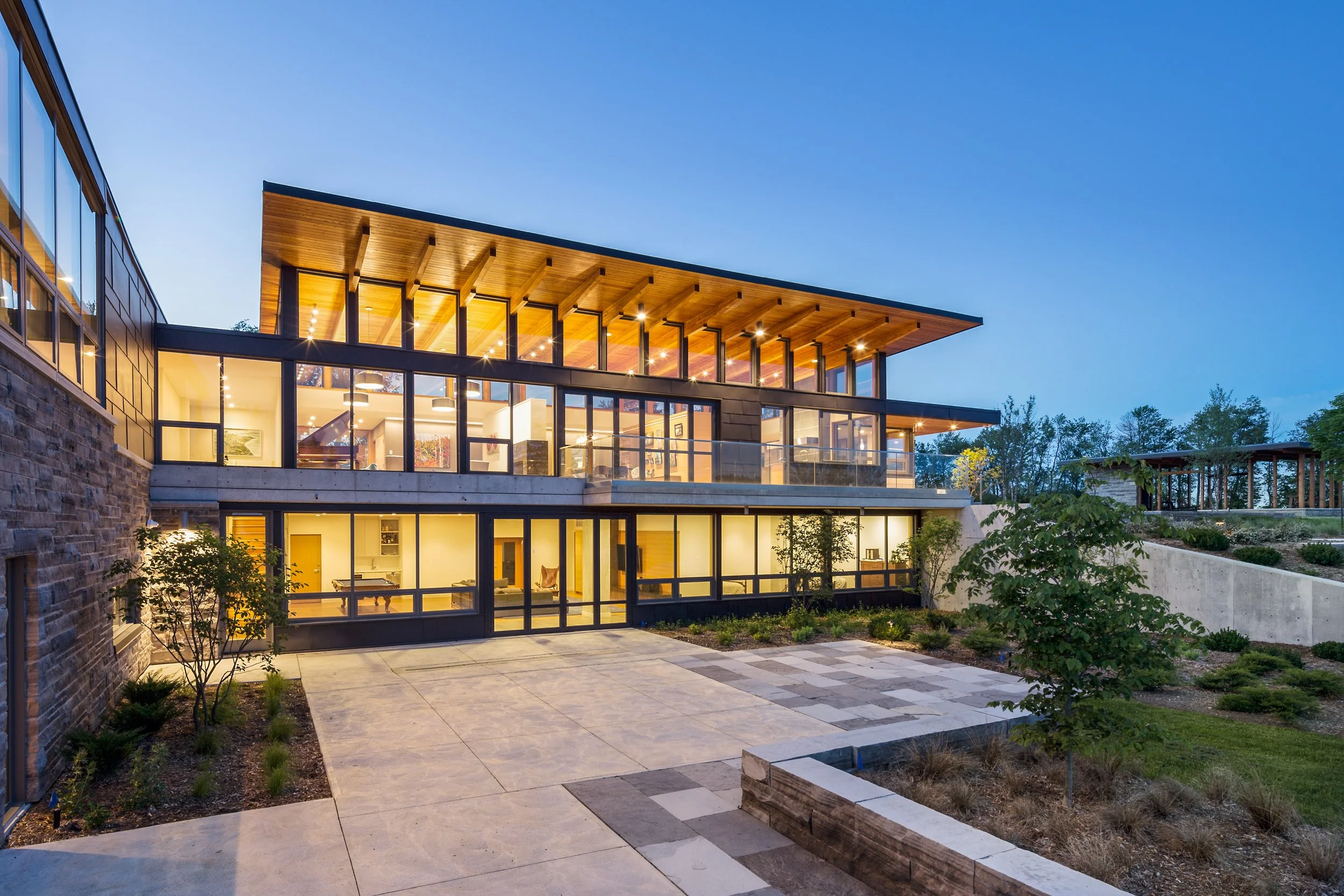 Modern multi-story house with large glass windows and wooden accents, illuminated at dusk, with a concrete patio and landscaped yard.