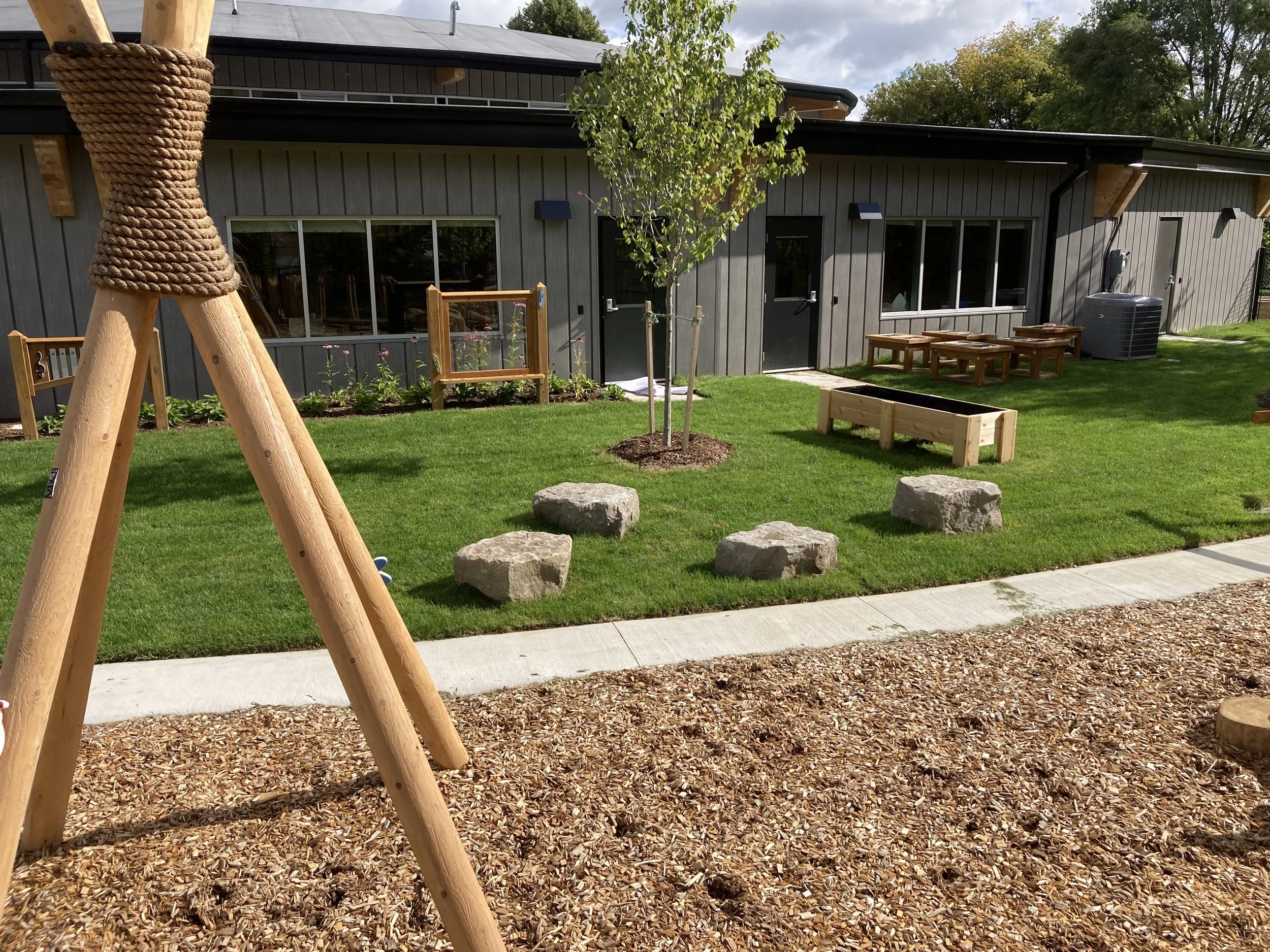 School playground with wooden swing set, grassy area, stone stepping stones, small trees, flower bed, and a gray building with large windows and outdoor furniture.