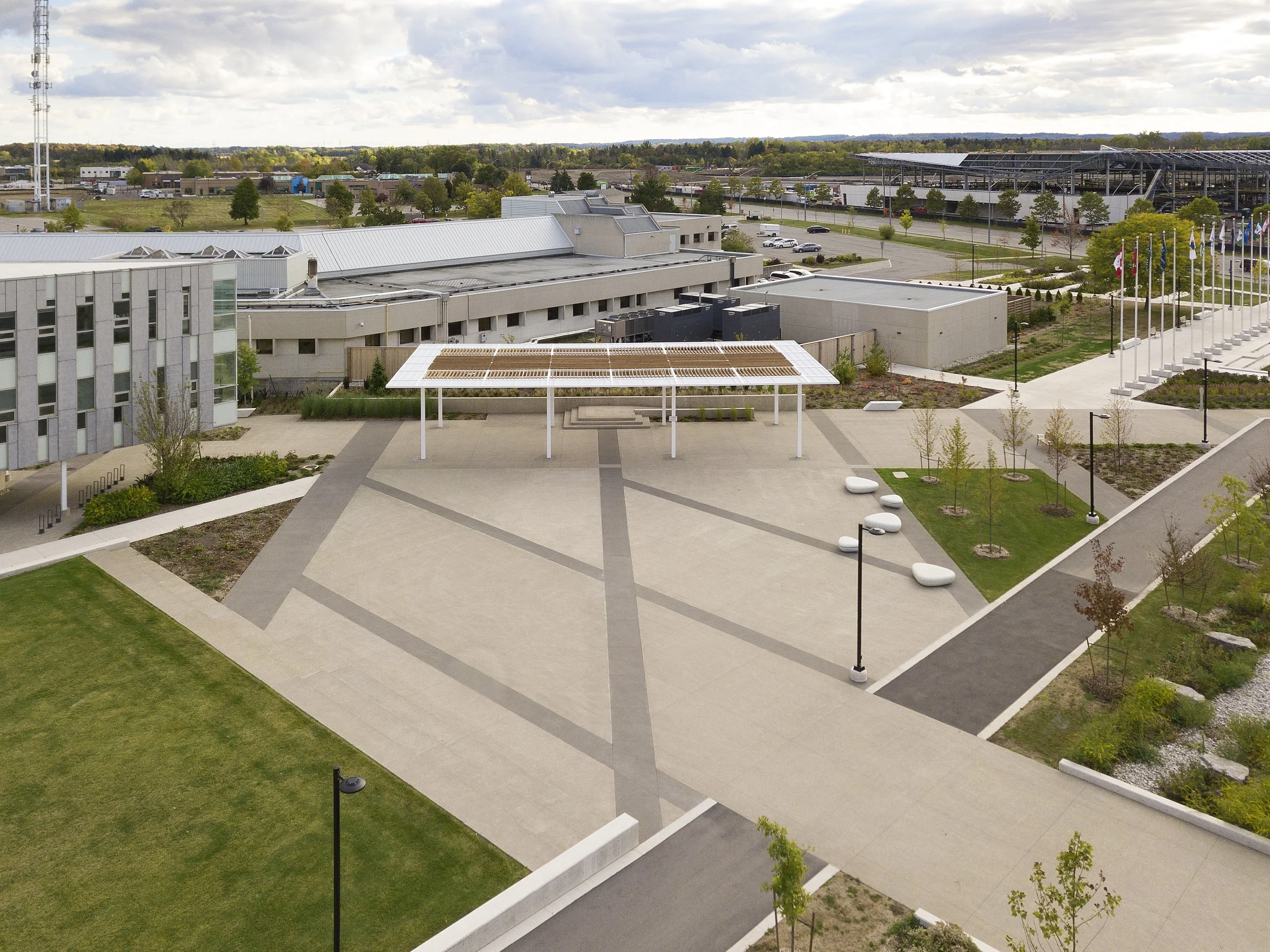 Aerial view of a modern outdoor plaza with pathways, green spaces, trees, benches, and a shaded structure, adjacent to a large complex with multiple buildings and a parking lot, with a cloudy sky overhead.
