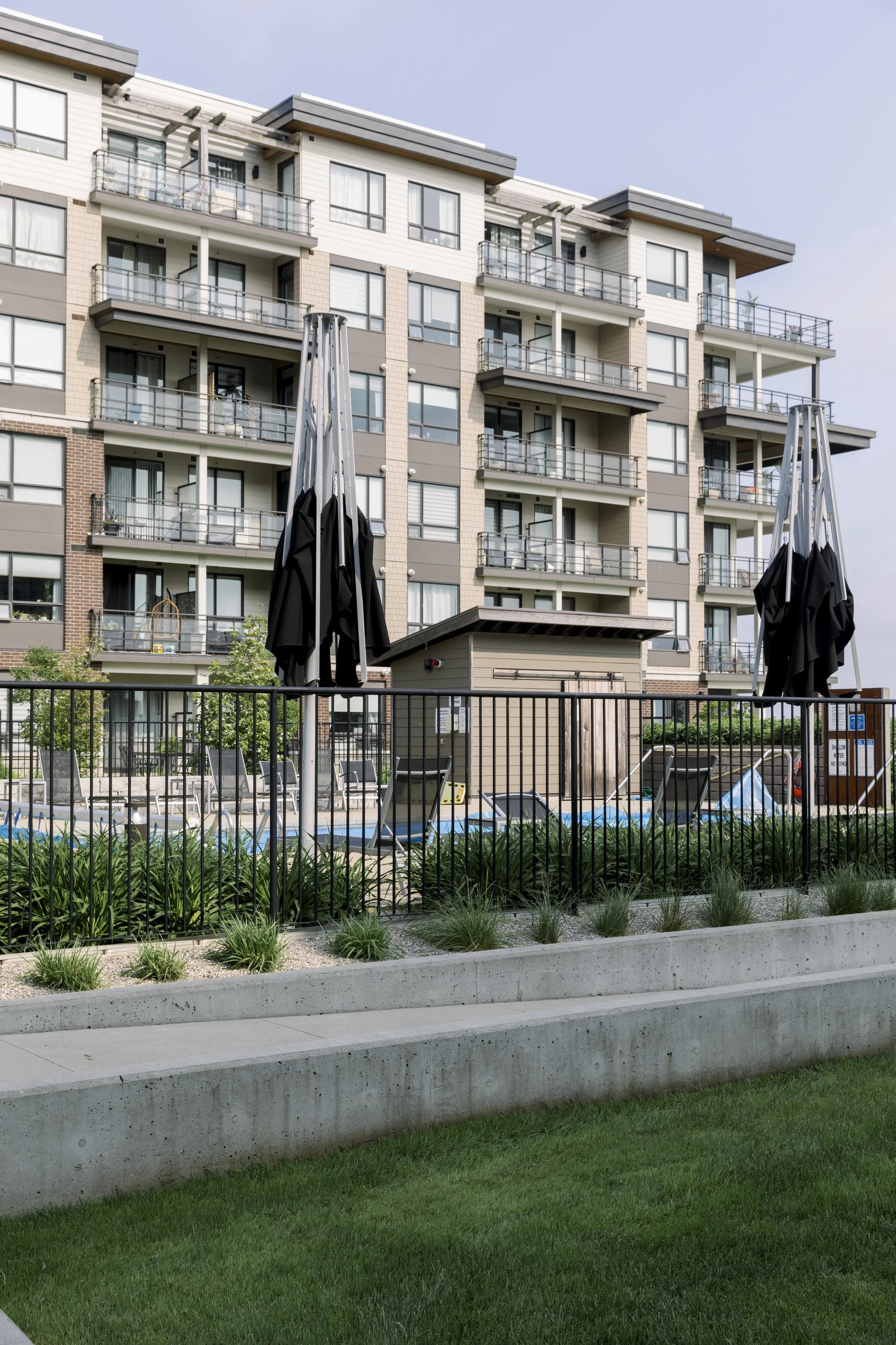 Exterior view of an apartment complex with a pool area, lounge chairs, and closed umbrellas, surrounded by a black metal fence and greenery.