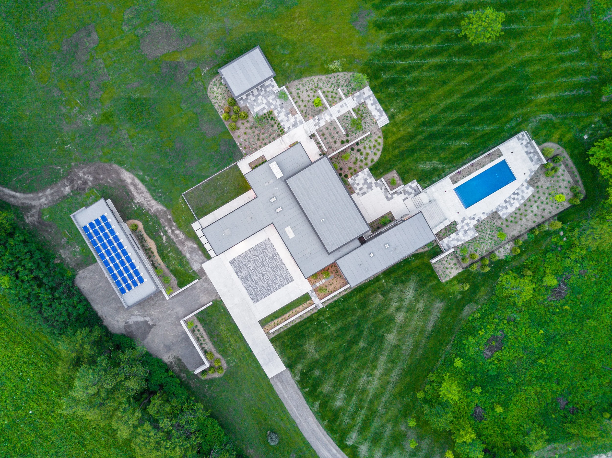 Aerial view of a modern house with a swimming pool, solar panels, and landscaped yard, surrounded by green grass and trees.