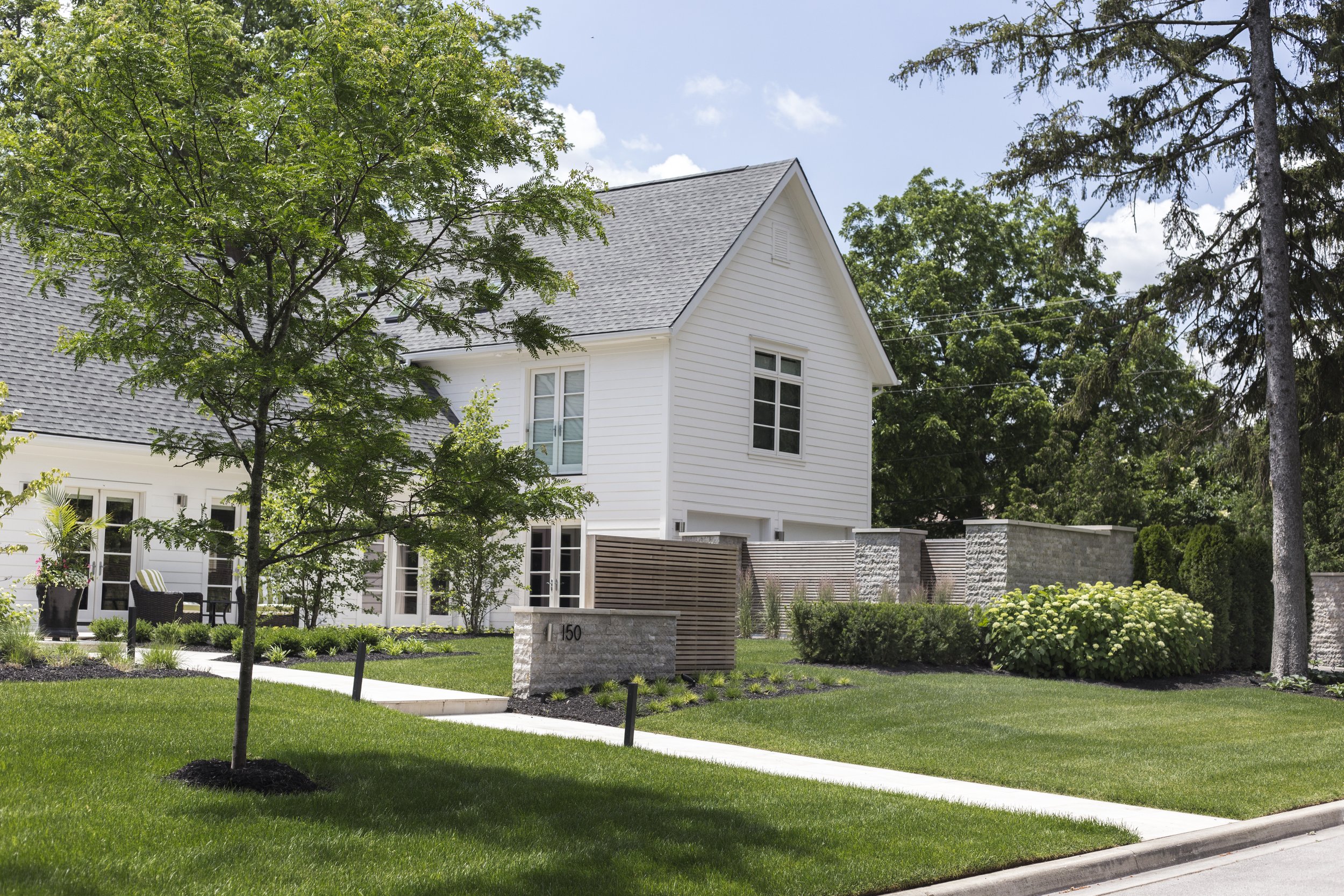 A modern white house with a gray roof, surrounded by a lush green lawn and trees, with a concrete sidewalk in front and a stone wall.