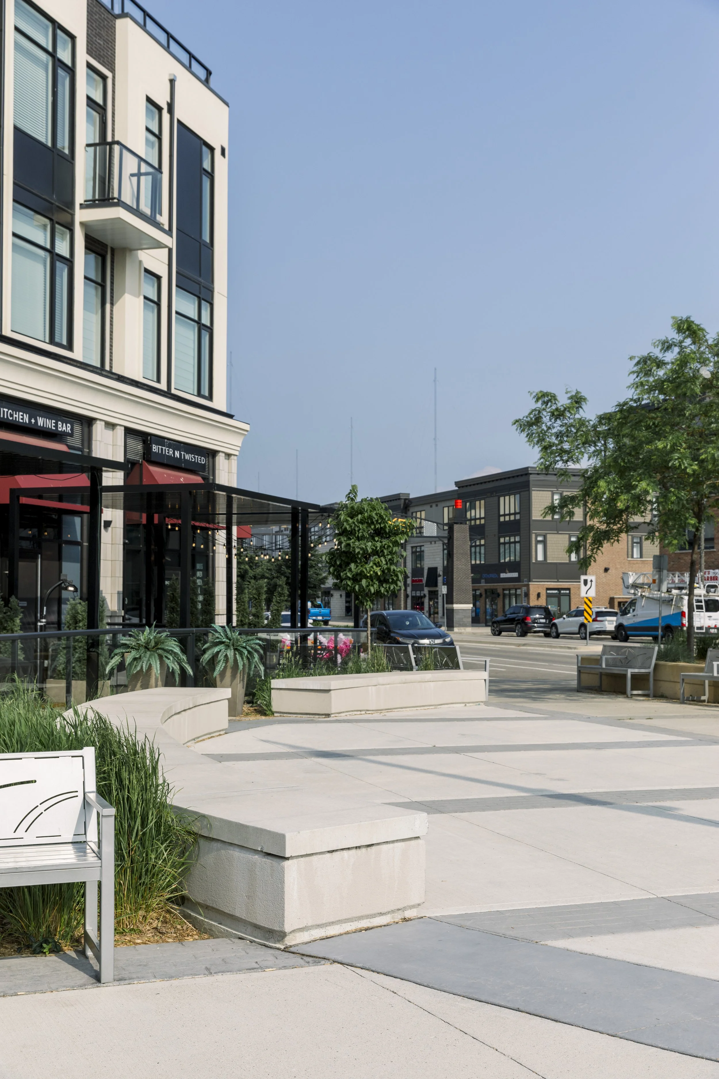 Empty urban sidewalk with benches, planters, and small trees, with buildings and cars in the background.