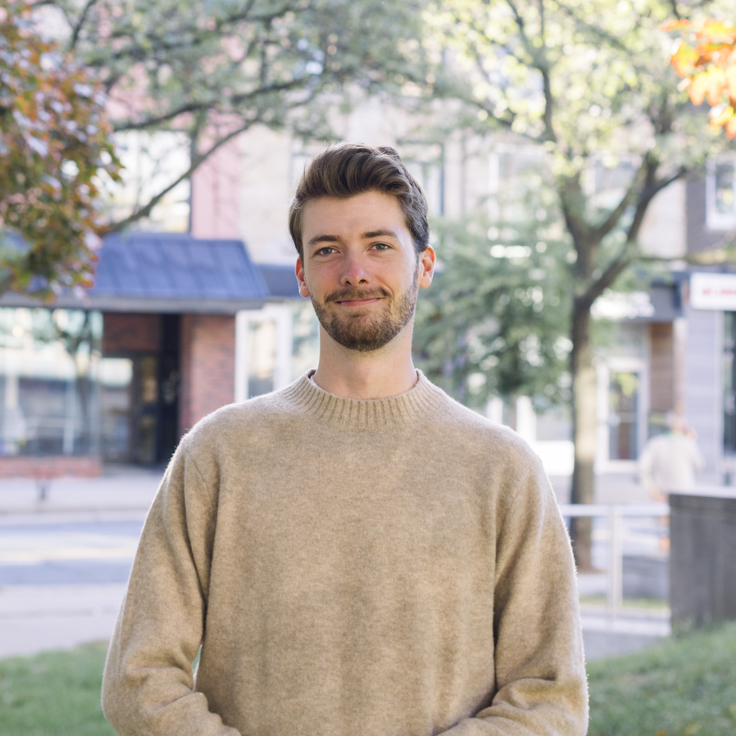 A young man with brown hair and a beard standing outdoors on a sunny day, wearing a beige sweater and smiling in front of trees and buildings.
